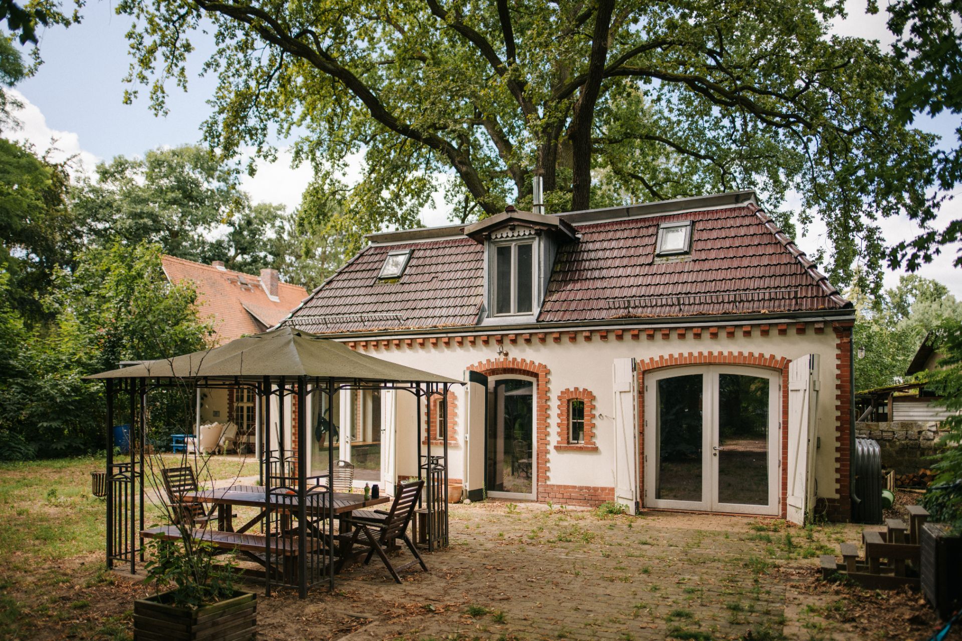 Kleines Haus mit Dachgauben und Terrasse, davor Pavillon und Gartenmöbel auf einem Naturgrundstück.