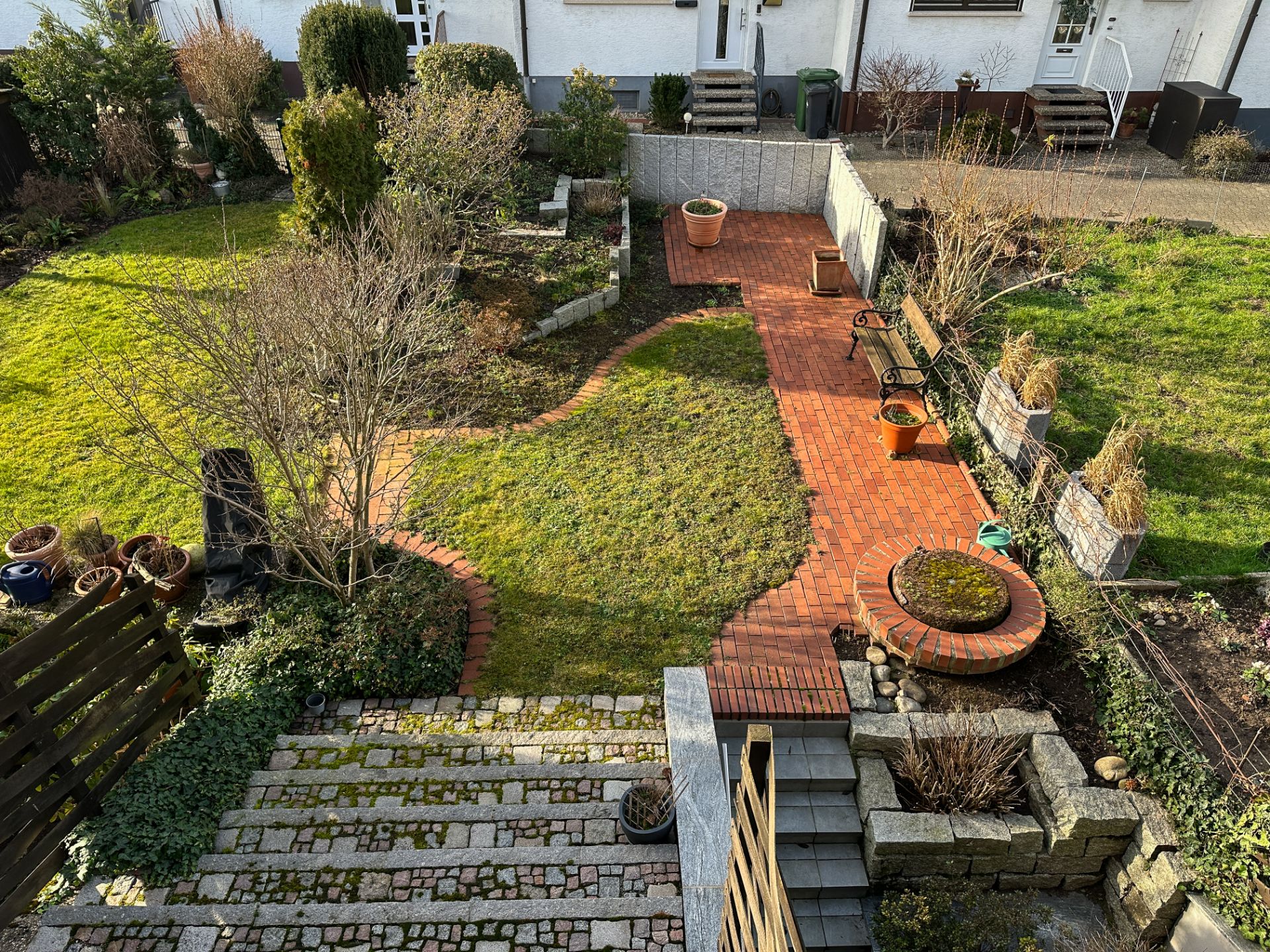 Gepflegter Garten mit Rasen, gepflastertem Weg, Treppen und mehreren Pflanzkübeln.