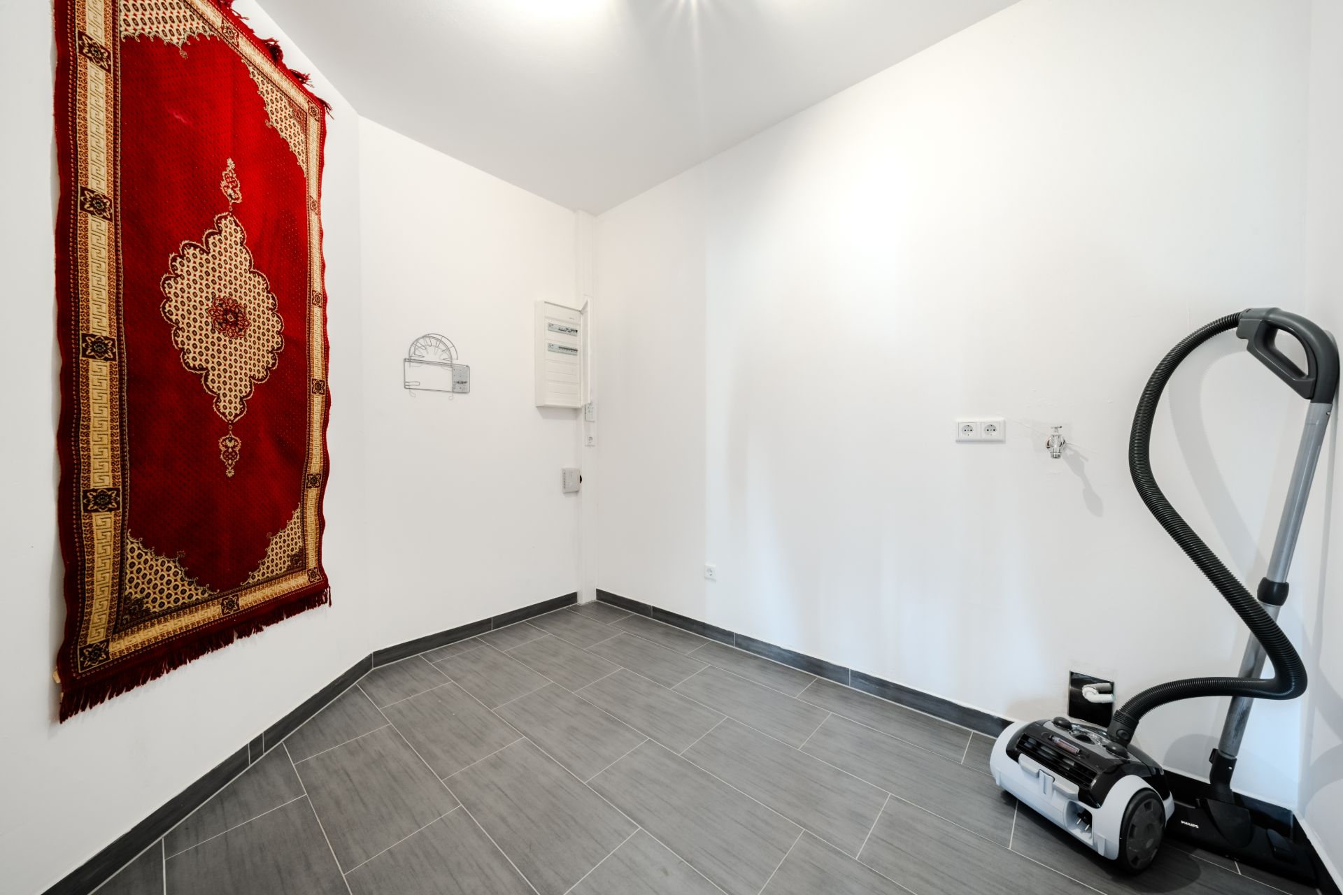 Kleiner Raum mit grauem Fliesenboden, Teppich an der Wand und Staubsauger in der Ecke.