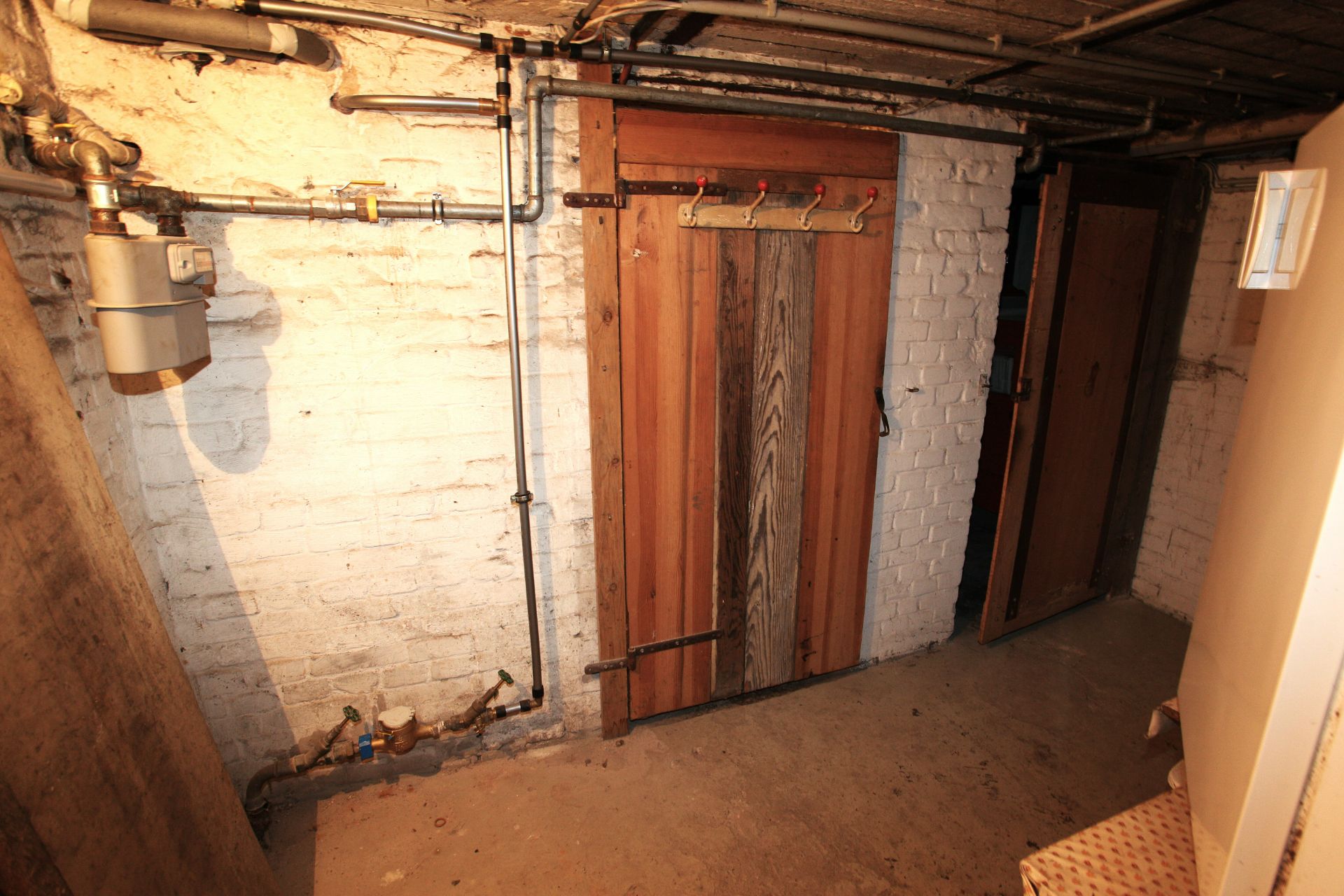 Kellerraum mit sichtbaren Leitungen, weißer Wand und einer Holztür.