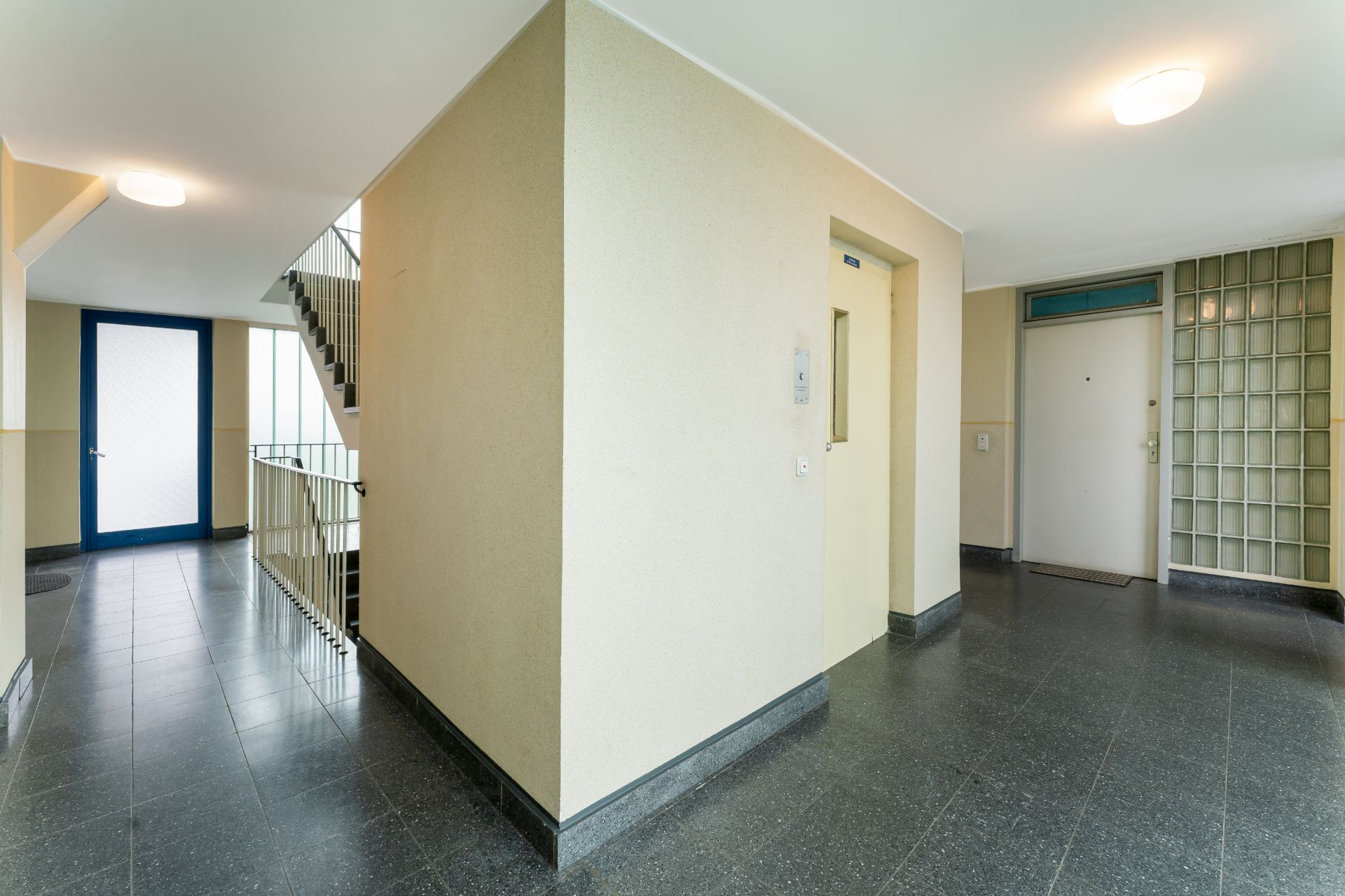 Heller Flur mit Treppenhaus, Aufzug und Wohnungs- sowie Glastür.