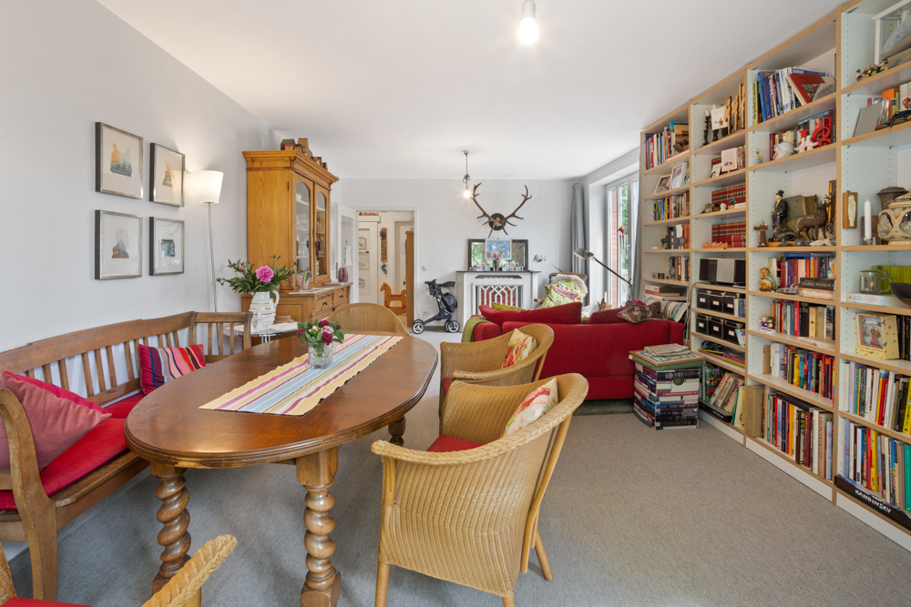 Wohnzimmer mit Esstisch, roten Sofas, Bücherregal und Holzschrank, dekoriert mit Blumen.