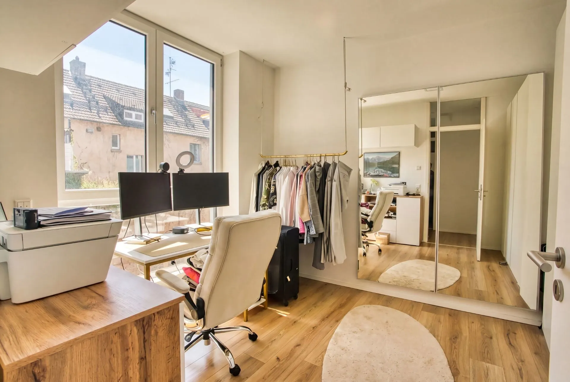 Helles Arbeitszimmer mit Schreibtisch, zwei Monitoren, Kleiderstange und großem Spiegel.
