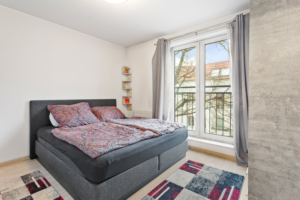 Schlafzimmer mit Doppelbett, bunter Bettwäsche, Teppich und bodentiefem Fenster mit Vorhängen.