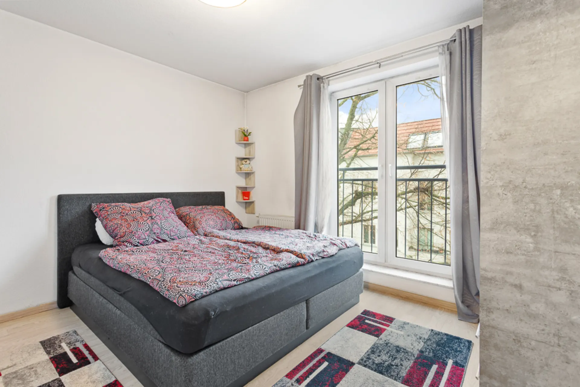 Schlafzimmer mit Doppelbett, bunter Bettwäsche, Teppich und bodentiefem Fenster mit Vorhängen.