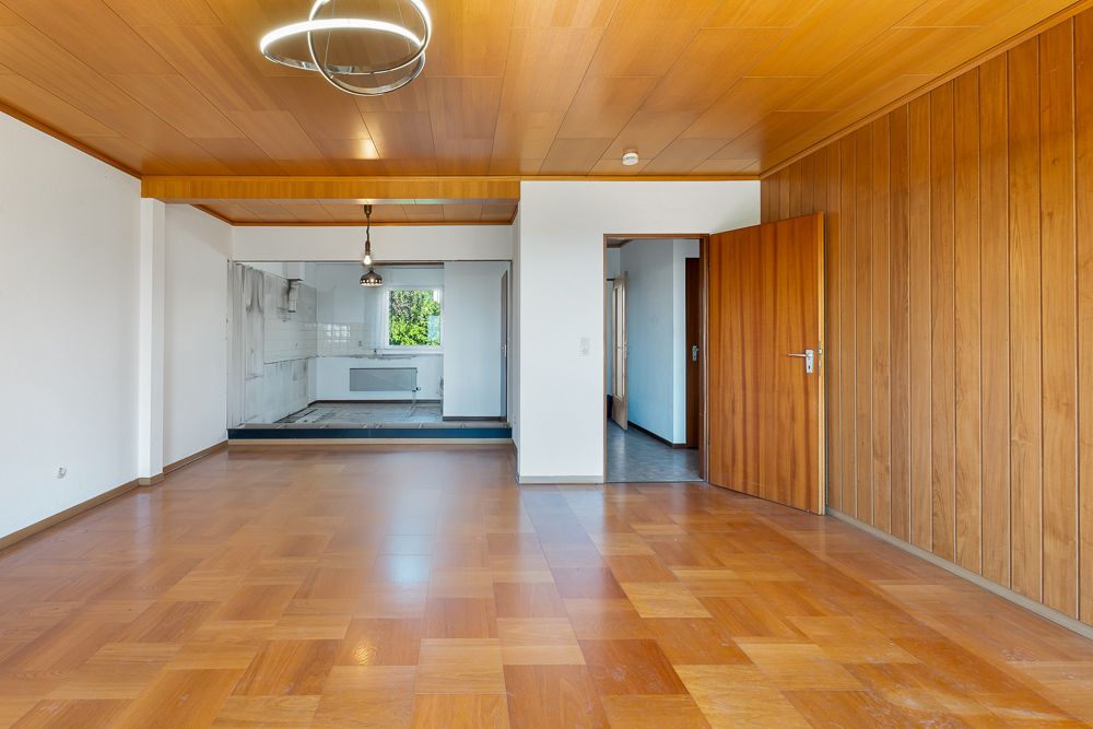 Helles Wohnzimmer mit Holzdecke, Holzboden, Sicht auf offene Küche und eine geöffnete Tür.