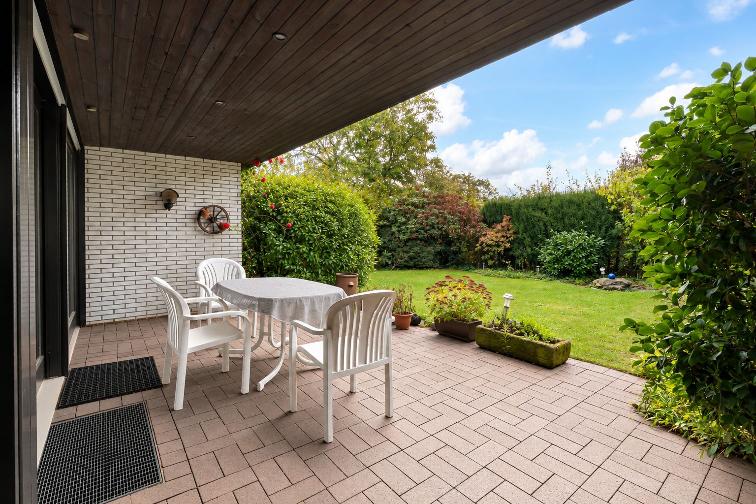Überdachte Terrasse mit Tisch, vier weißen Stühlen und Blick auf einen Garten mit Grünpflanzen.