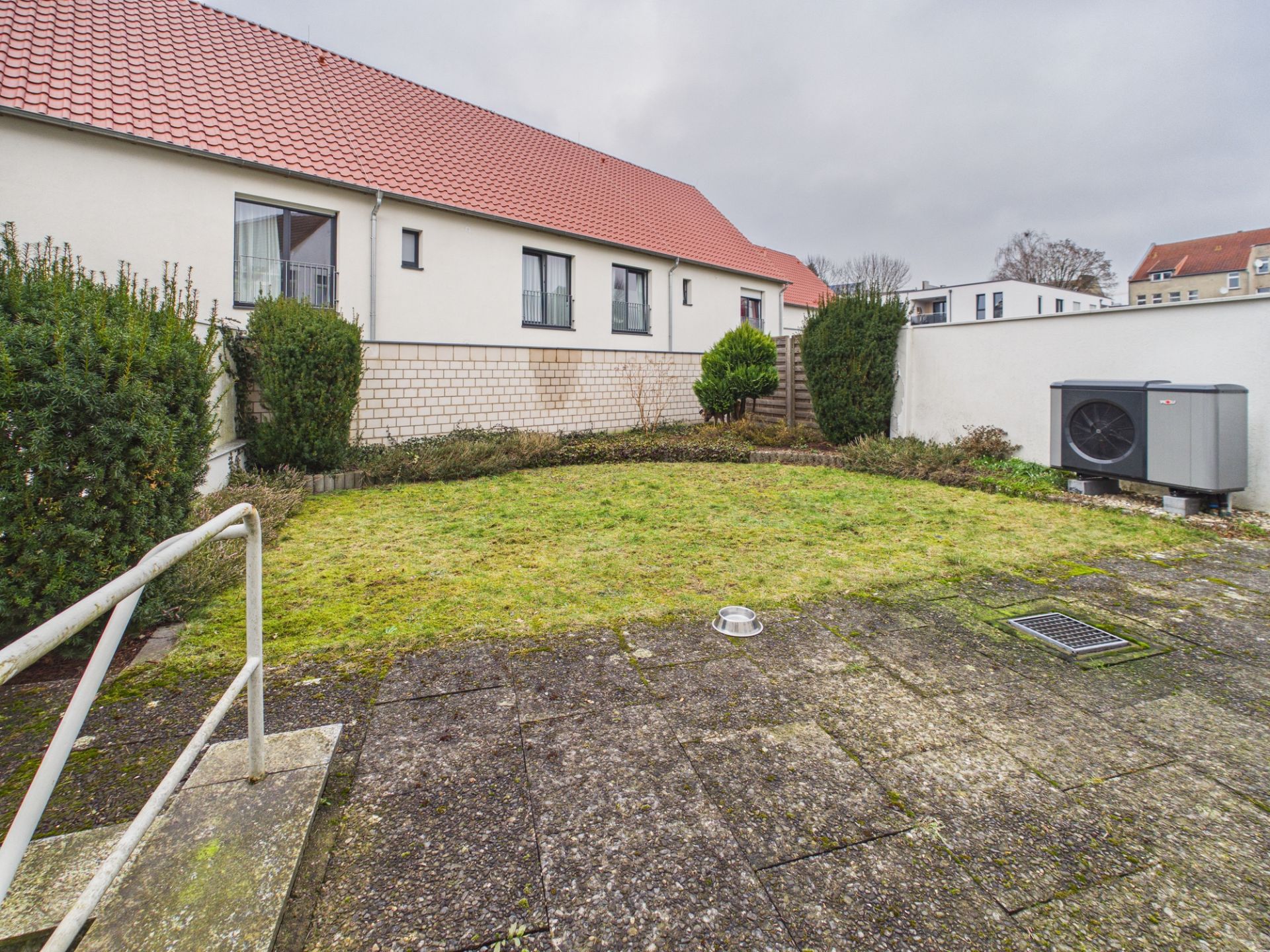 Gepflasterte Terrasse mit Grünfläche, Büschen und Heizungsanlage neben einem Haus mit rotem Dach.