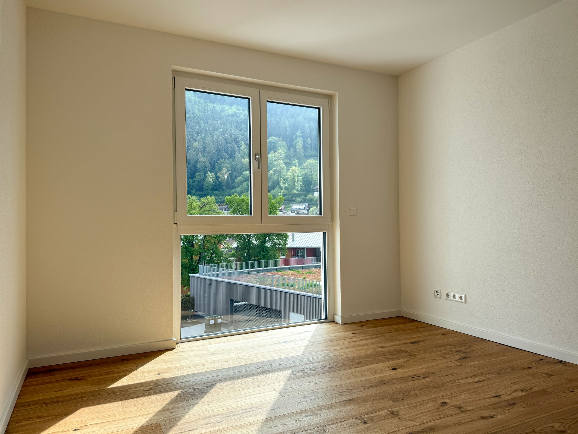 Helles leeres Zimmer mit Holzfußboden und großem Fenster mit Blick ins Grüne.