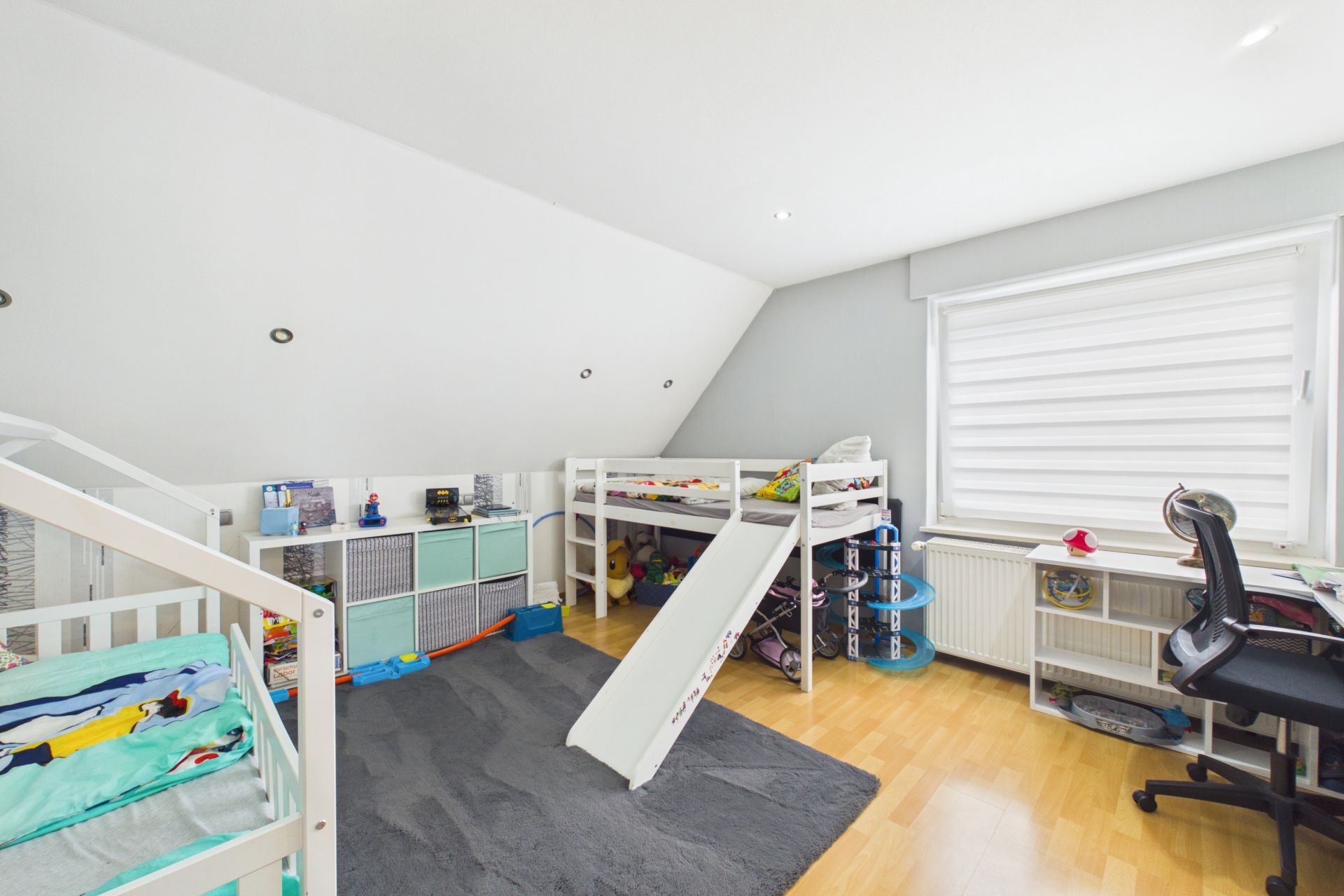 Kinderzimmer mit zwei Betten, Rutsche, Schreibtisch und großem Fenster.