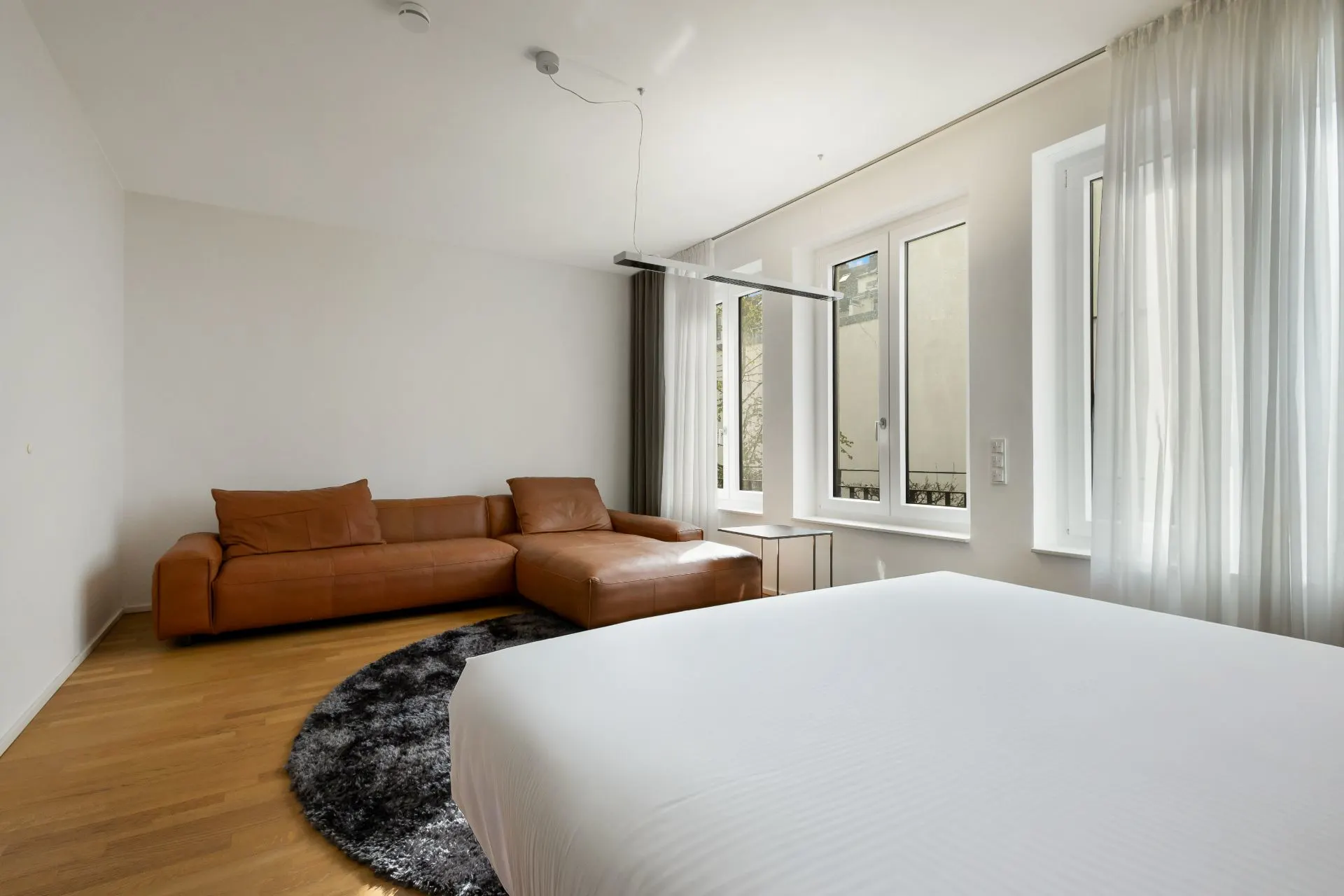 Helles Schlafzimmer mit braunem Sofa, großem Bett und drei Fenstern mit weißen Vorhängen.