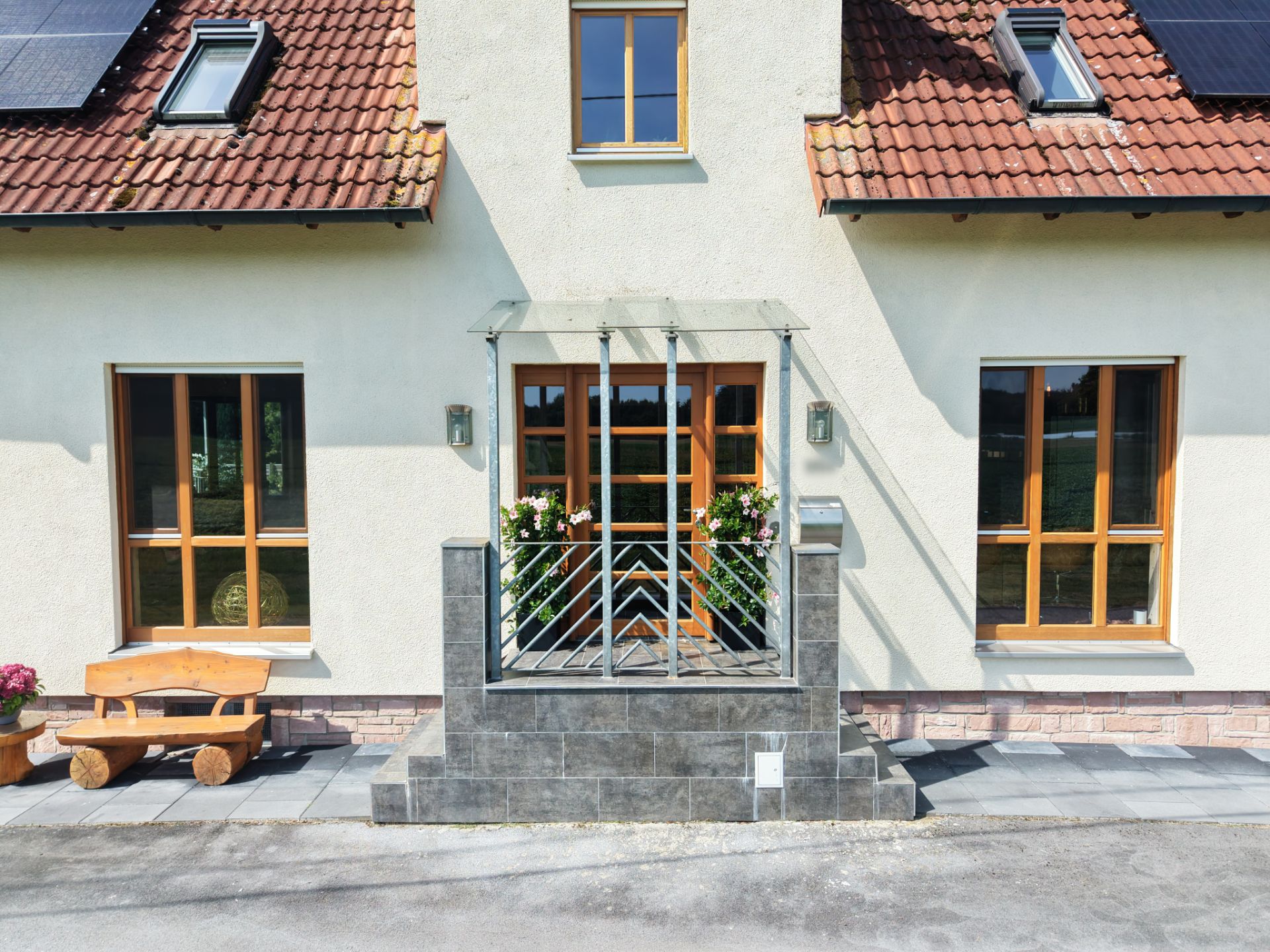 Eingang eines Wohnhauses mit Treppe, Geländer, Blumen und Holzbänken vor großen Fenstern.