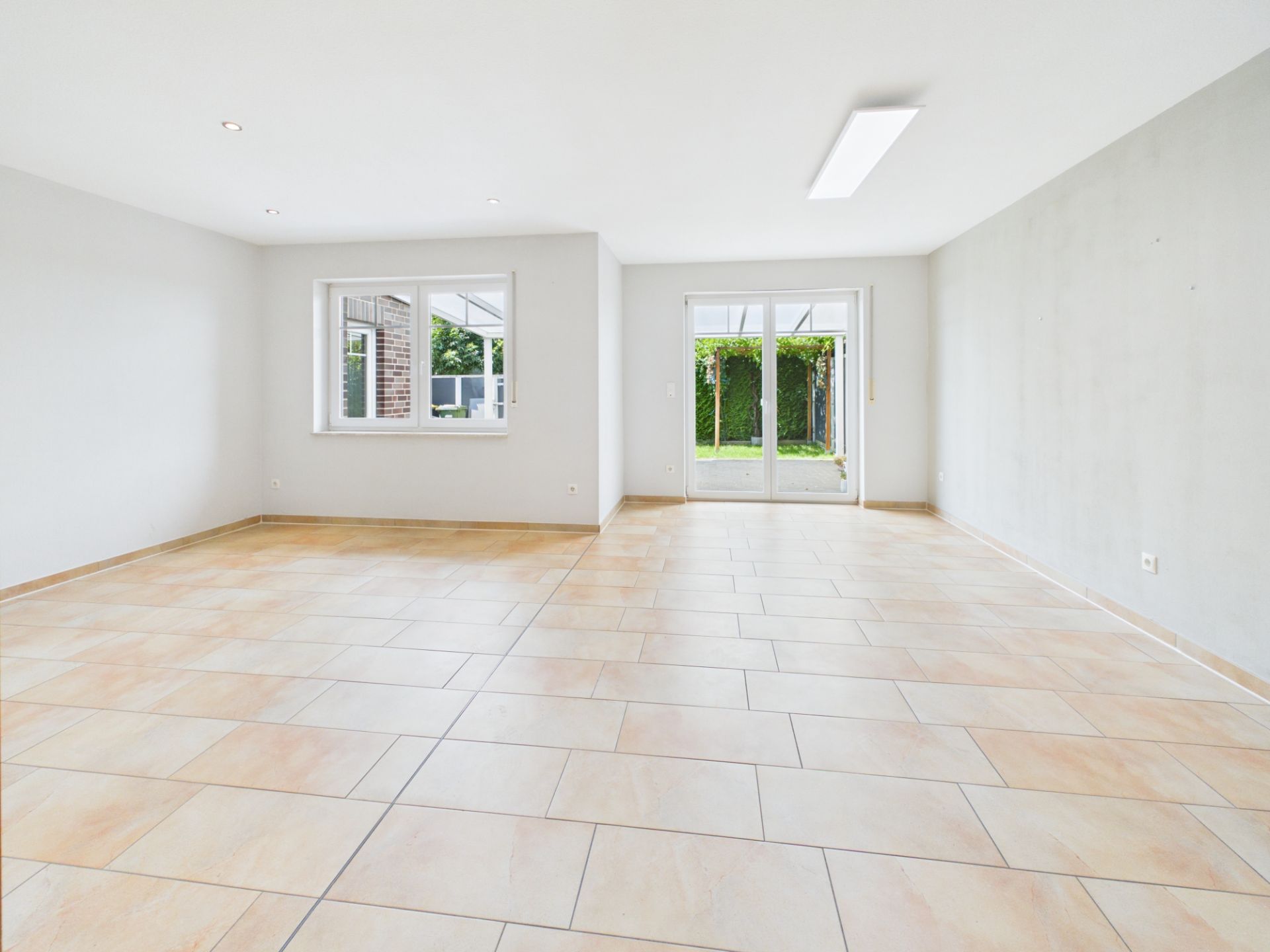 Heller, leerer Raum mit großen Fenstern und Terrassentür, gefliestem Boden und Blick in den Garten.