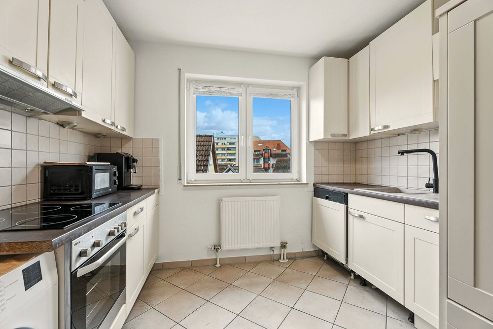 Helle Küche mit weißen Schränken, Backofen und Fenster mit Blick auf Wohngebäude.