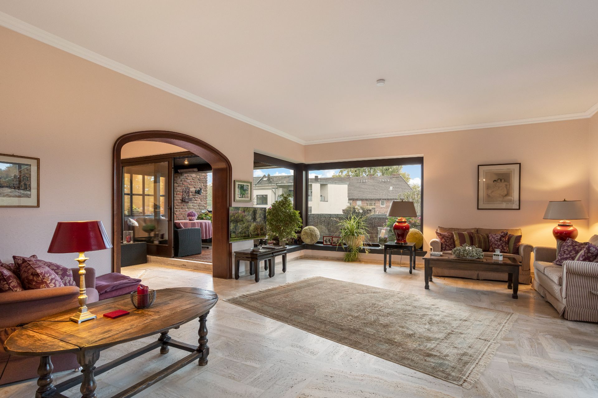 Helles Wohnzimmer mit Sofas, Teppich, großen Fenstern und Pflanzen, angrenzender Sitzbereich sichtbar.