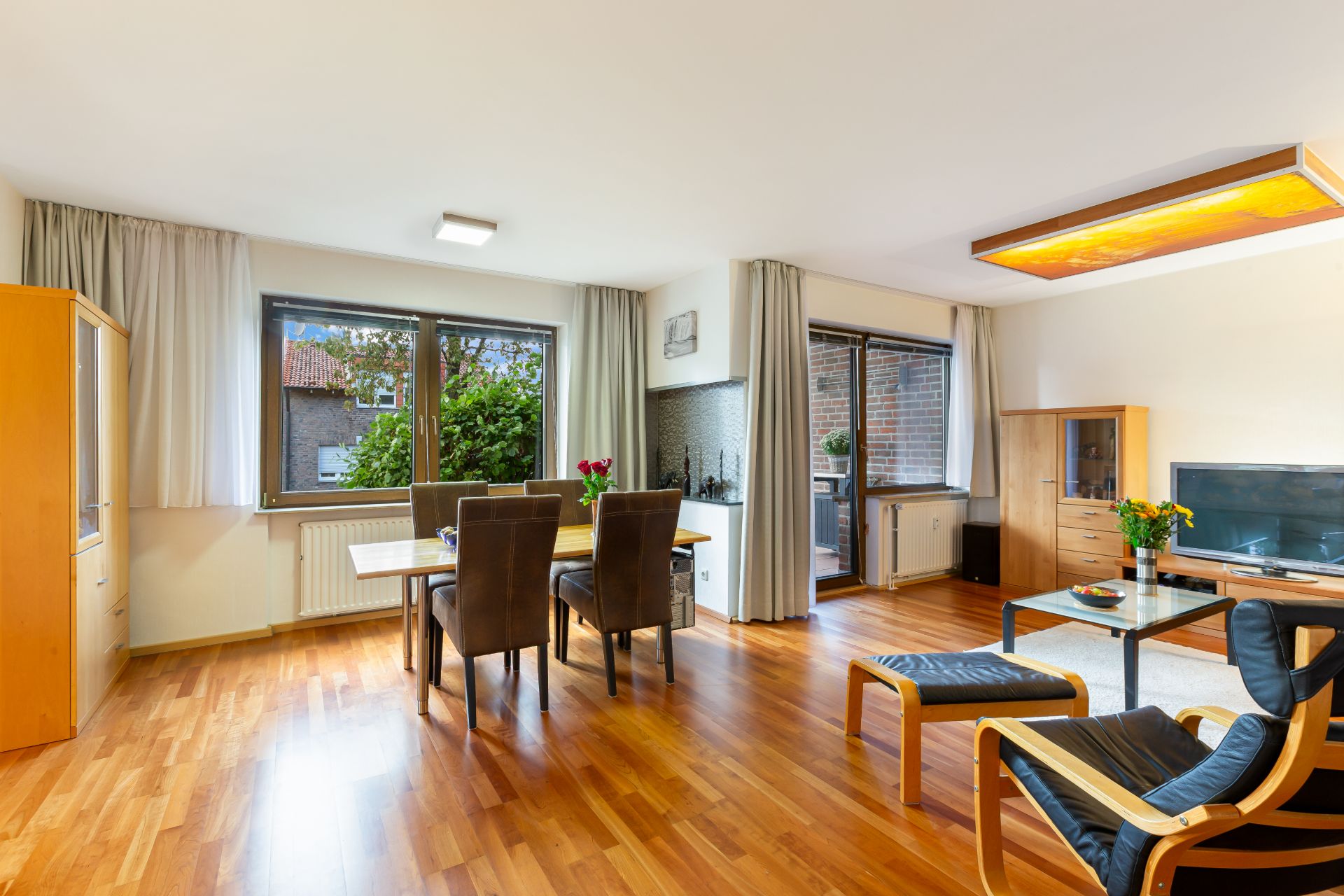 Helles Wohnzimmer mit Holzboden, Esstisch, Stühlen, Sofa, TV und großen Fenstern.