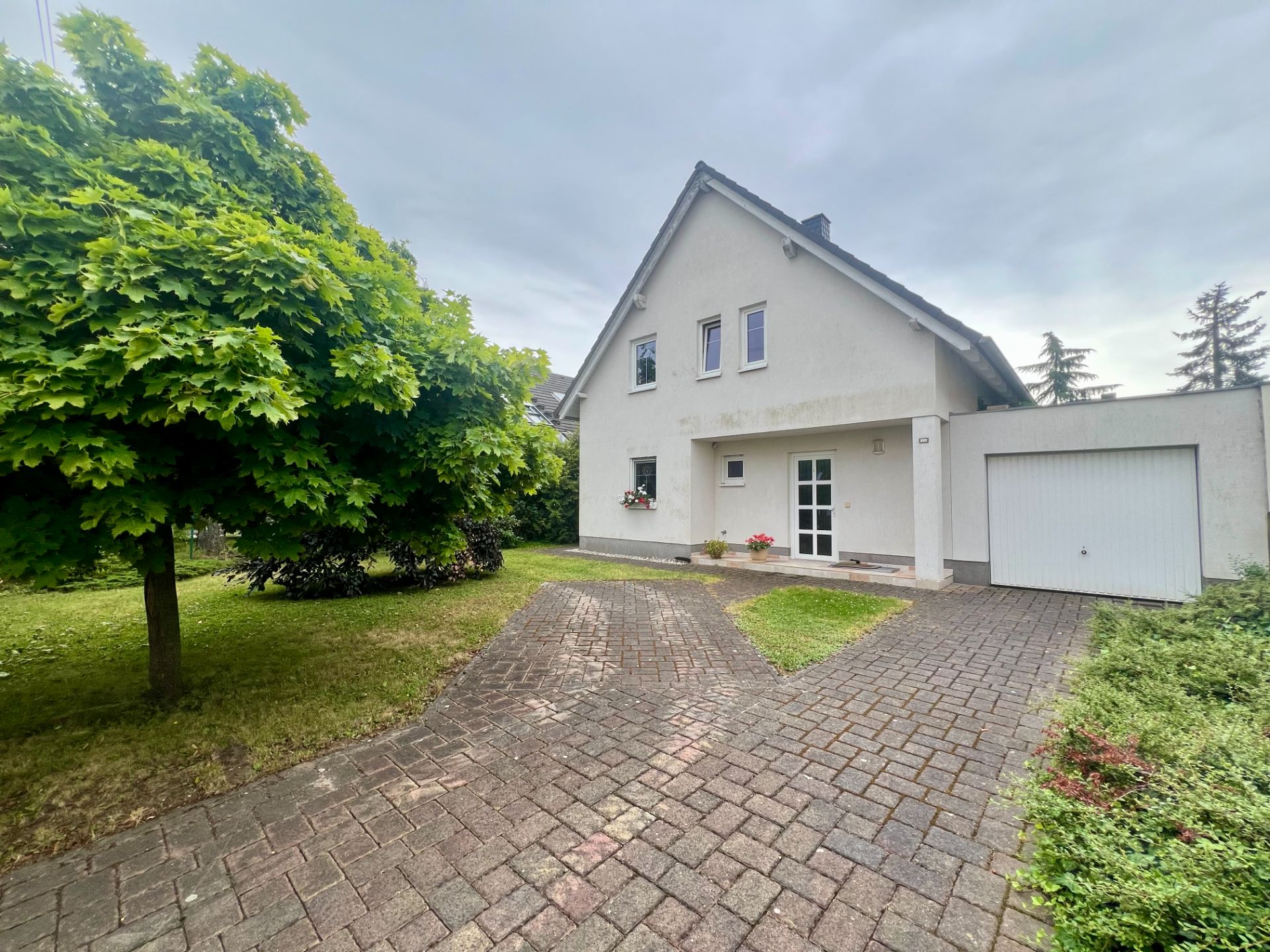 Einfamilienhaus mit Garage, gepflasterter Einfahrt und großem Baum vorne.