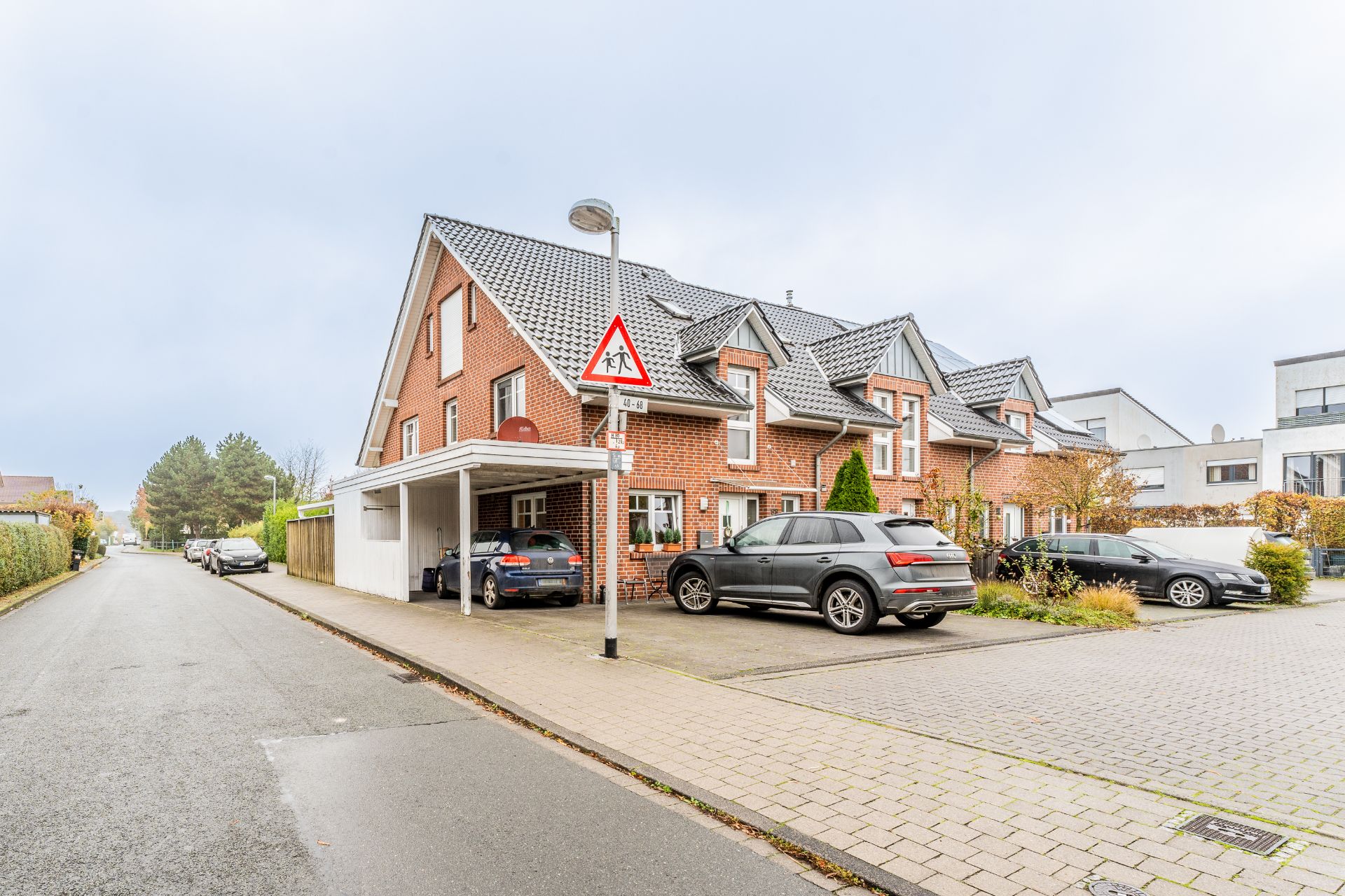 Backstein-Doppelhaushälfte mit Carport und geparkten Autos an einer ruhigen Wohnstraße.