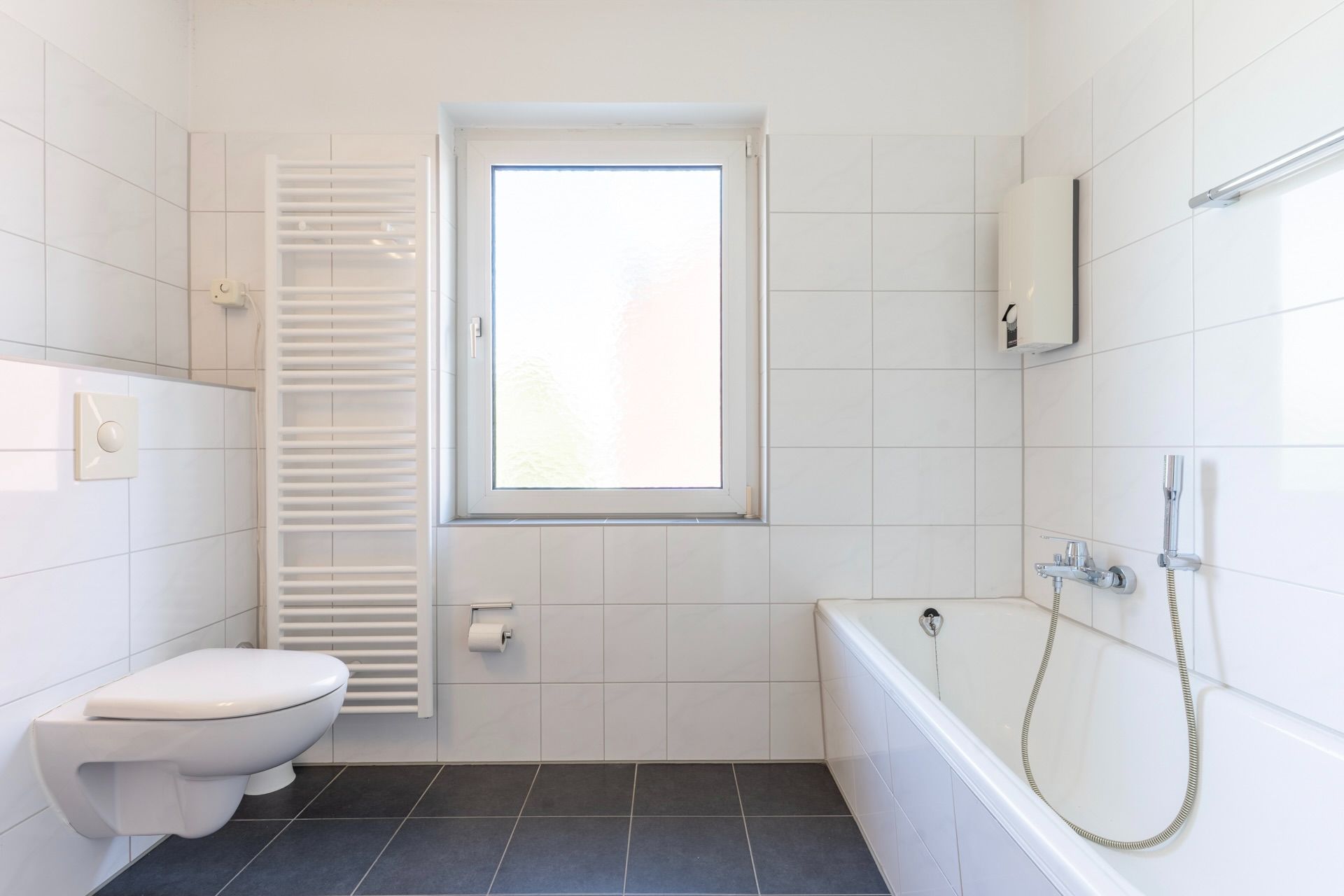 Helles Badezimmer mit Badewanne, Wandtoilette, Fenster und Handtuchheizkörper.