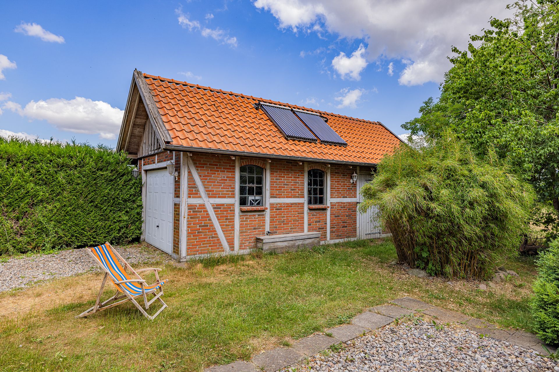 Backsteinanbau mit rotem Ziegeldach, Solarpanel, Liegestuhl und Garten im Vordergrund.