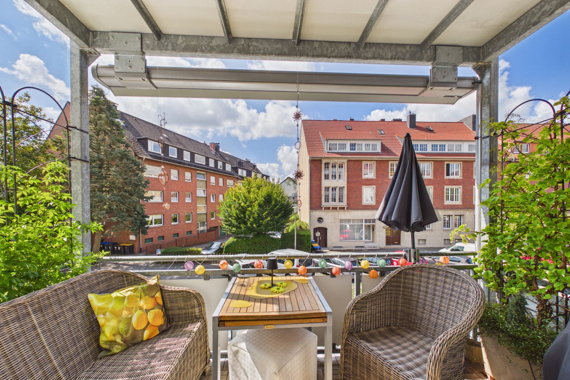 Balkon mit zwei Korbstühlen, Tisch, Pflanzen und Blick auf Wohnhäuser.