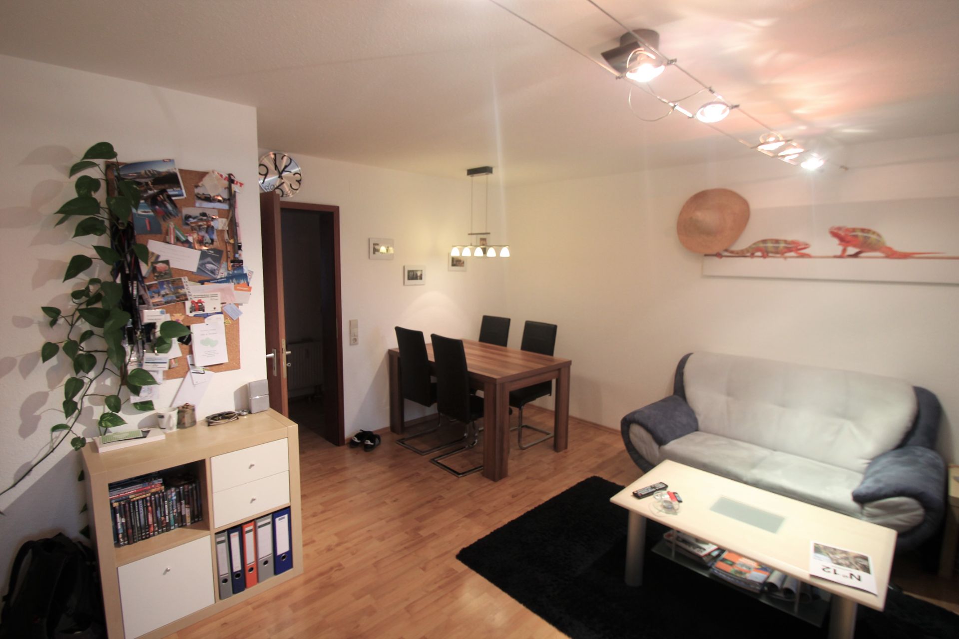 Wohnzimmer mit Sofa, Esstisch mit vier Stühlen, Regal und Pinnwand an der Wand.