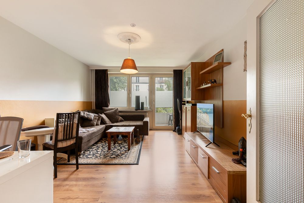 Wohnzimmer mit Sofa, Couchtisch, Fernseher, Regal und großem Fenster mit Blick ins Freie.