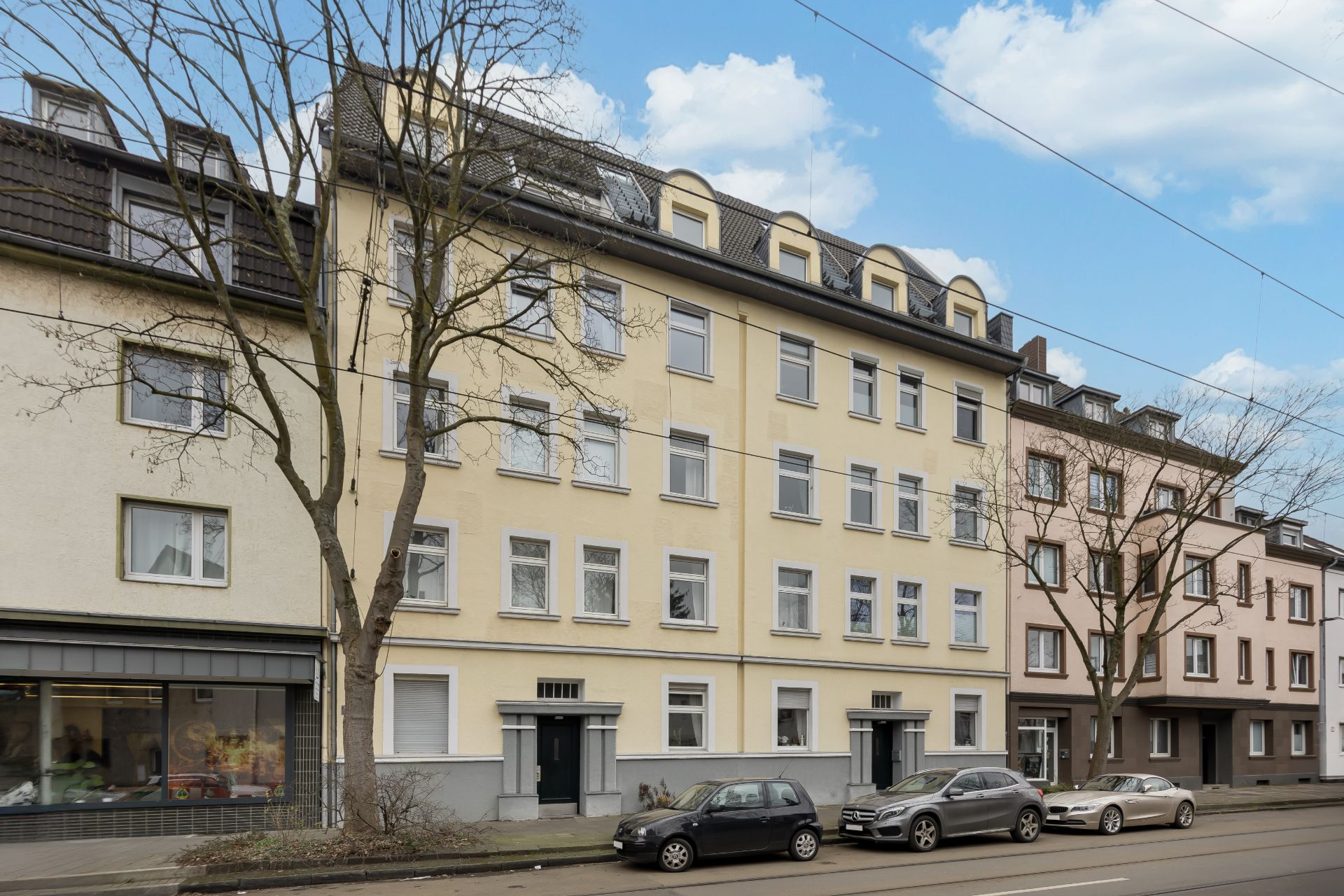 Mehrstöckiges Wohnhaus mit gelber Fassade, parkende Autos und kahle Bäume davor.