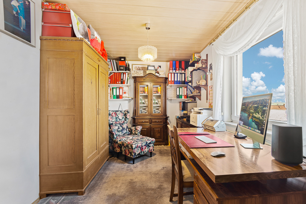 Helles Arbeitszimmer mit Schreibtisch, Computer, Schrank, Regal und Fenster mit Blick nach draußen.