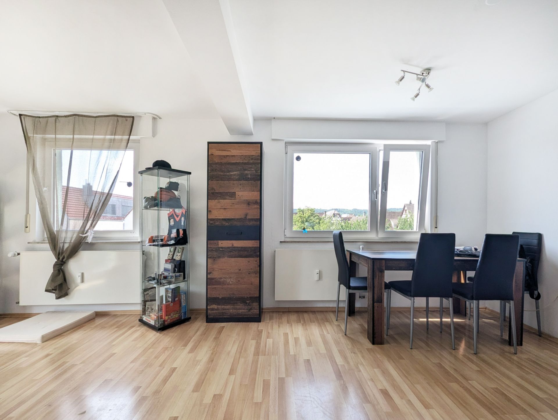 Helles Zimmer mit Esstisch, vier Stühlen, Vitrine, Schrank und zwei Fenstern.
