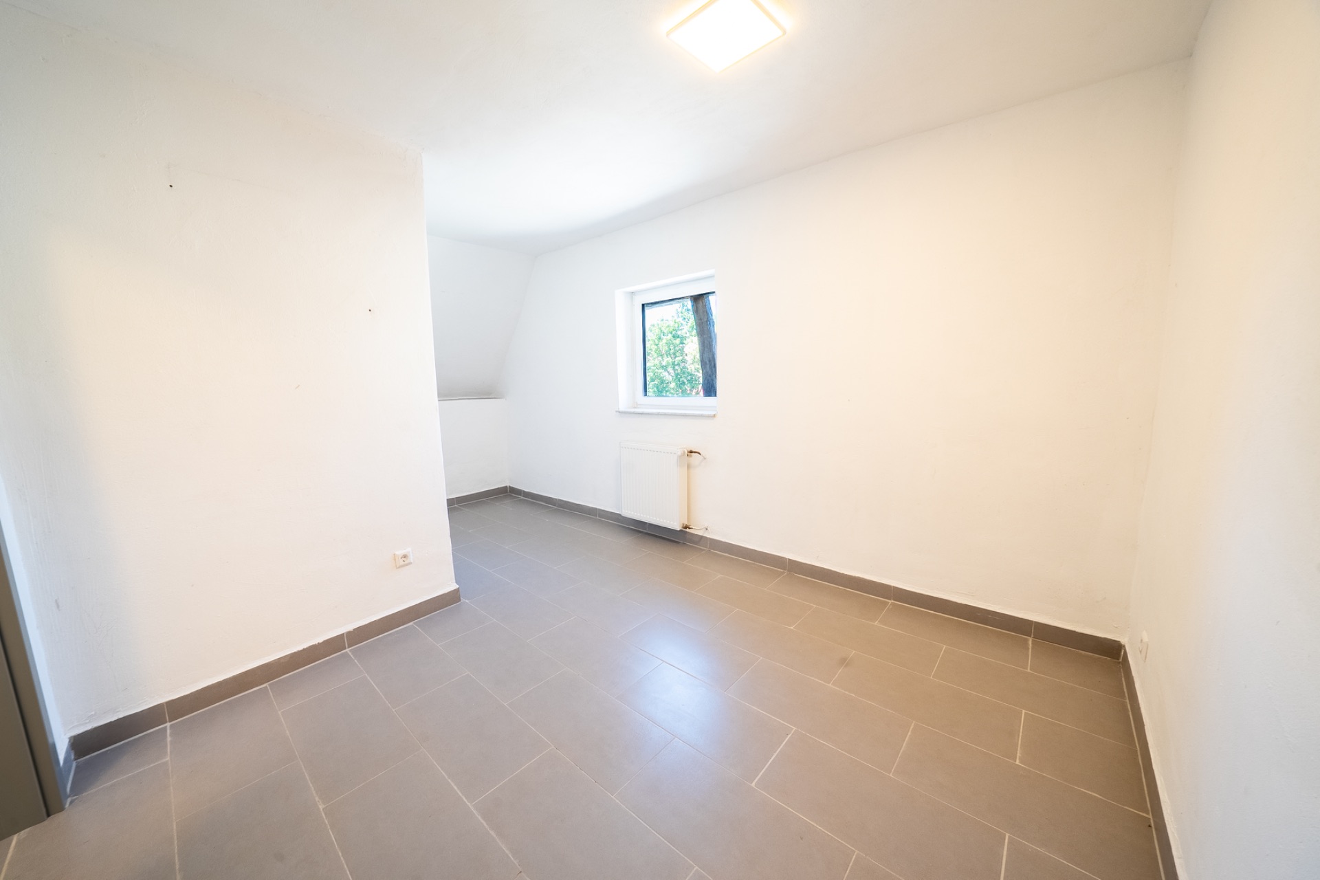 Leerer, heller Raum mit grauem Fliesenboden und einem Fenster an der Wand.