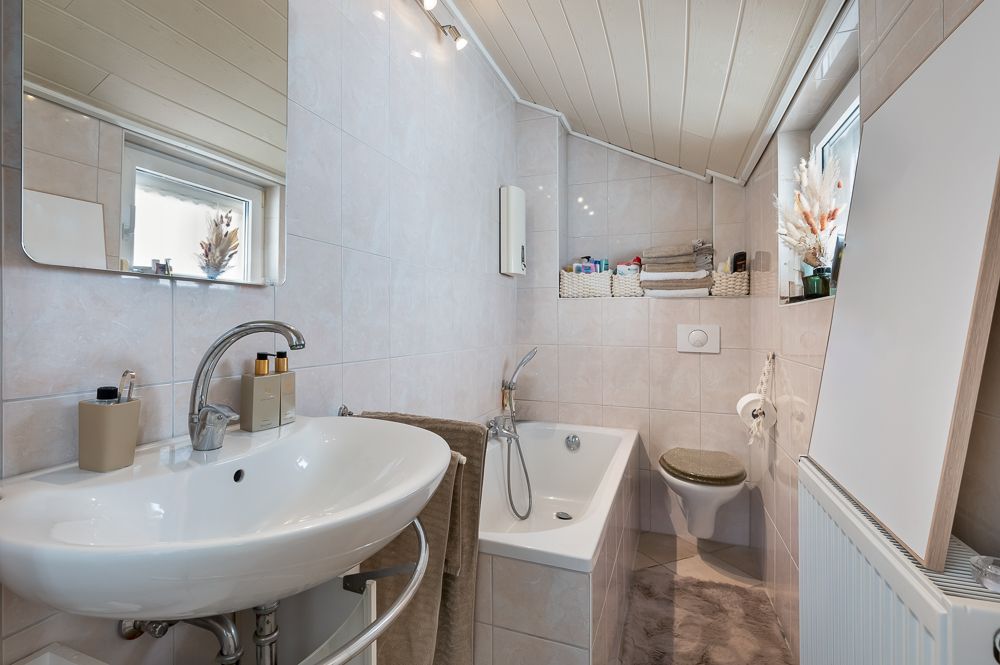 Helles Badezimmer mit Waschbecken, Badewanne, Toilette und zwei Fenstern unter Dachschräge.
