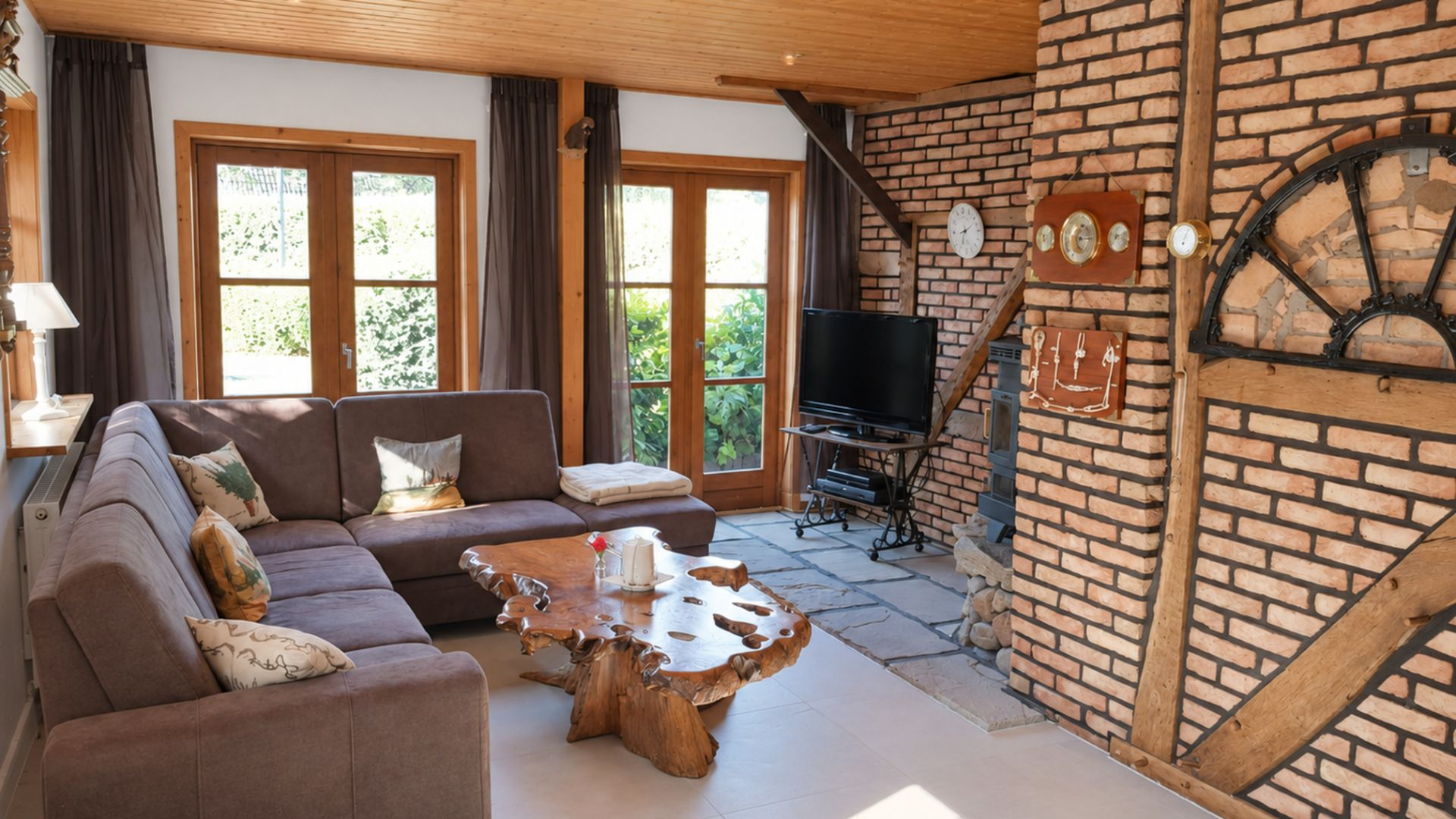 Wohnzimmer mit Ecksofa, Holztisch, TV, Backsteinwand und großen Fenstern mit Blick ins Grüne.