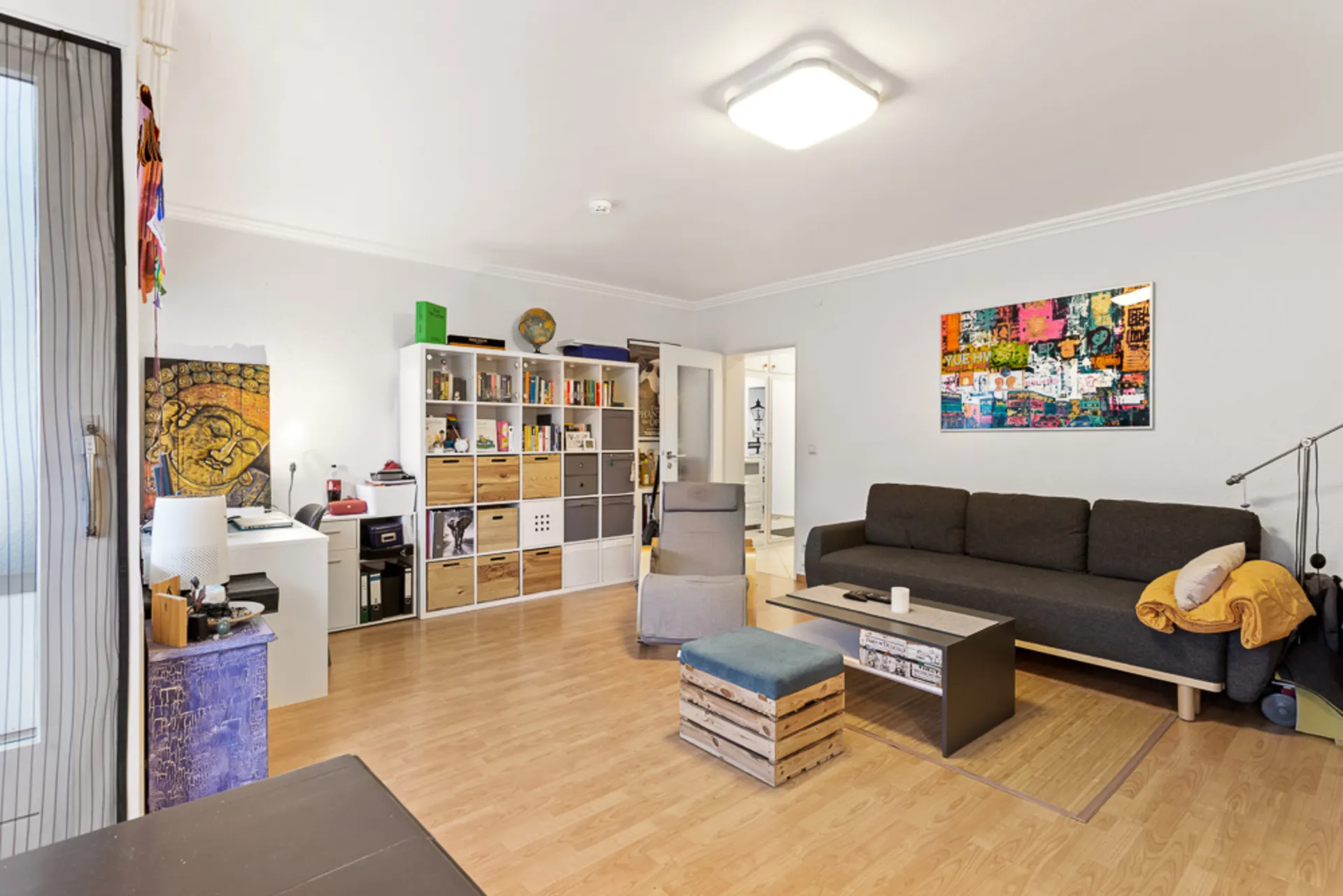 Wohnzimmer mit schwarzem Sofa, Regal, Schreibtisch und farbenfrohem Wandbild.