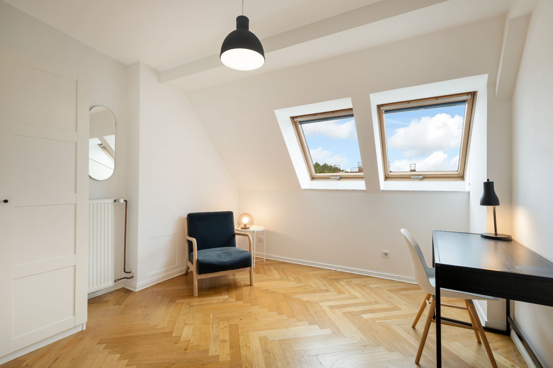 Helles Zimmer mit Sessel, Schreibtisch, Stuhl und zwei Dachfenstern.
