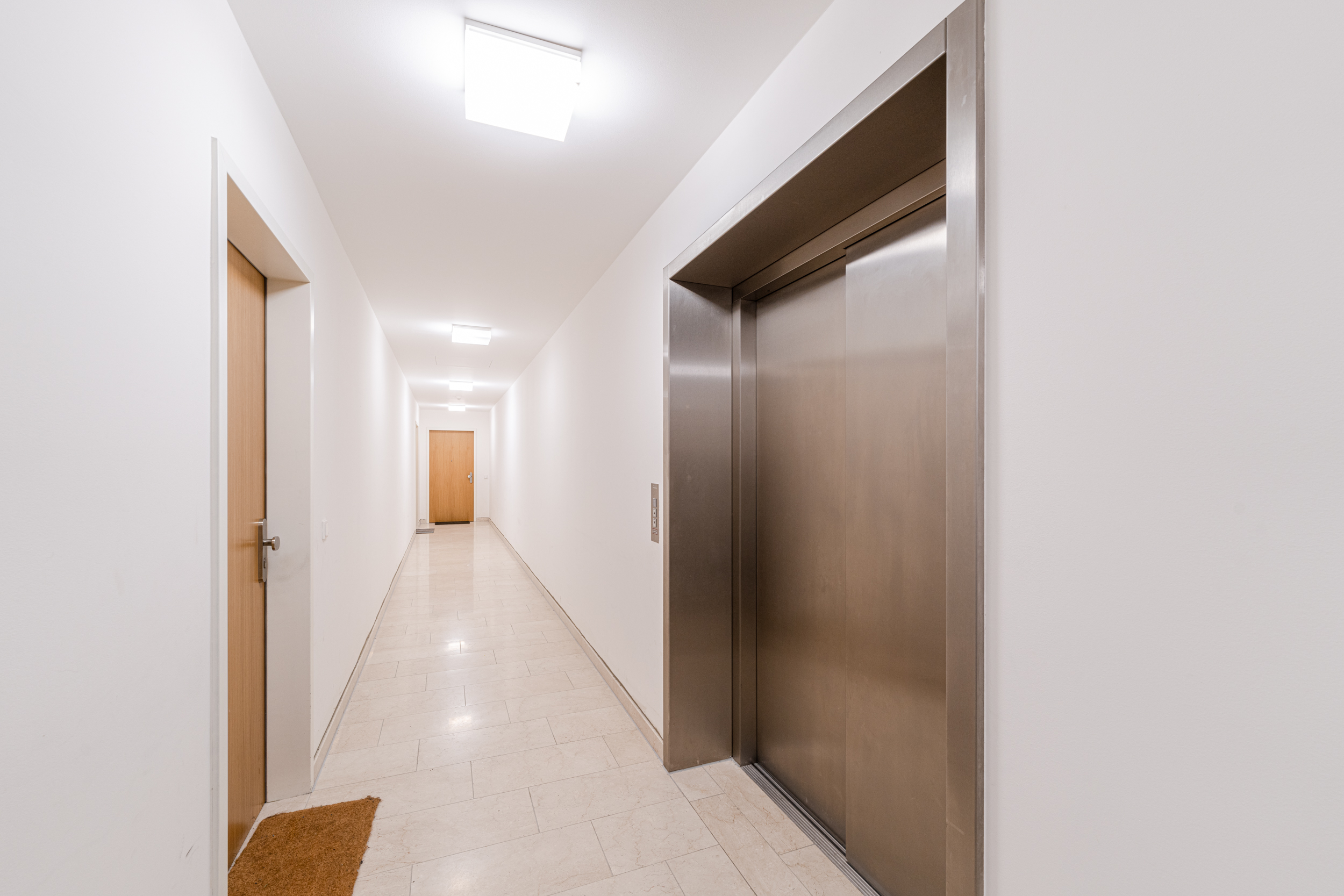 Heller Flur mit Fahrstuhl, Holz- und Wohnungstüren, Fliesenboden und Deckenbeleuchtung.