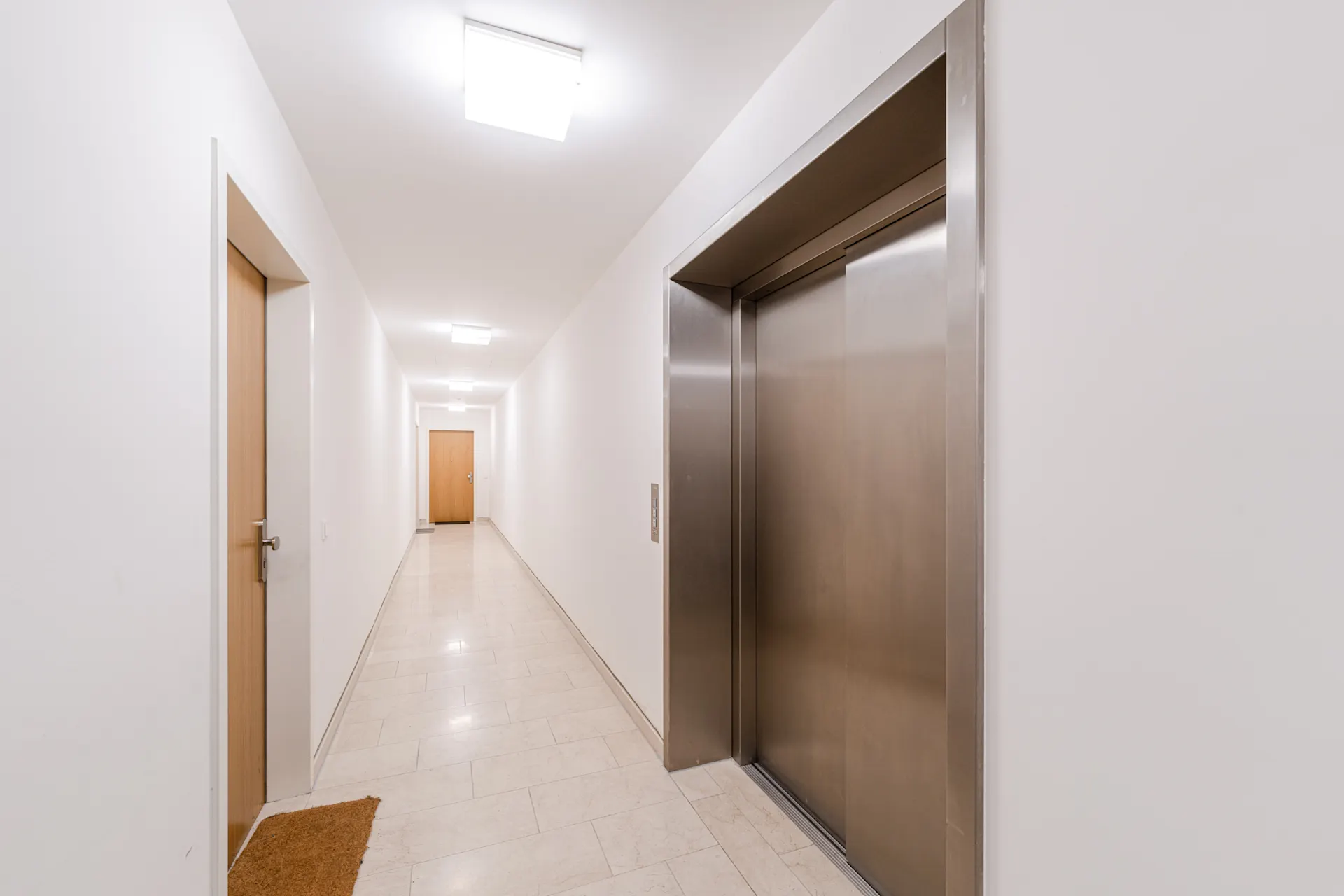 Heller Flur mit Fahrstuhl, Holz- und Wohnungstüren, Fliesenboden und Deckenbeleuchtung.