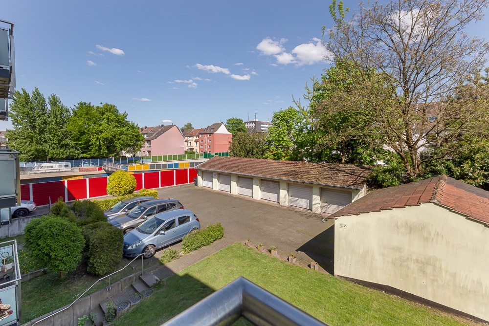 Blick auf Parkplatz mit Autos und Garagen, im Hintergrund Wohnhäuser und Bäume.