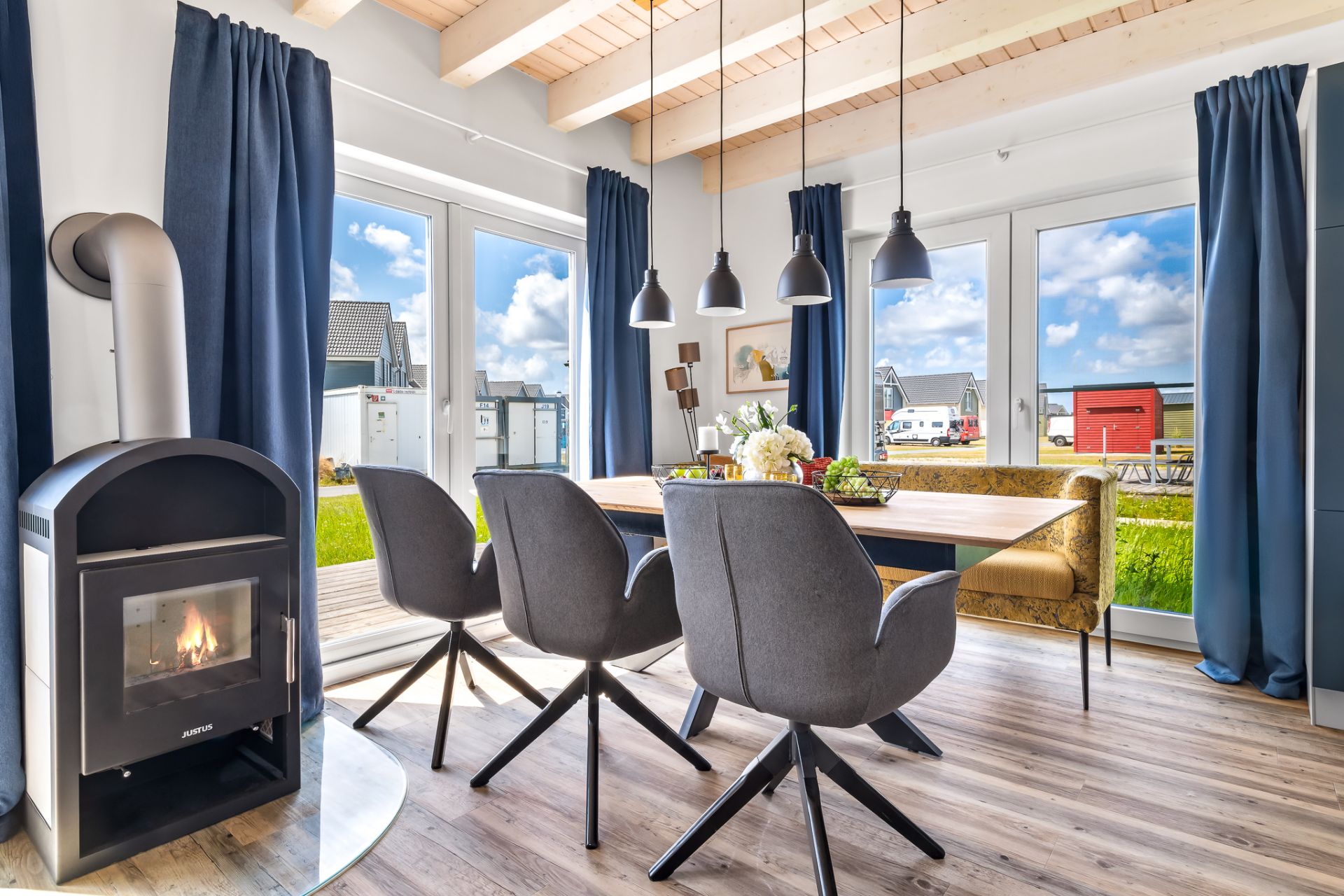 Heller Essbereich mit Kamin, Holztisch, grauen Stühlen, Sofa und großen Fenstern zum Garten.