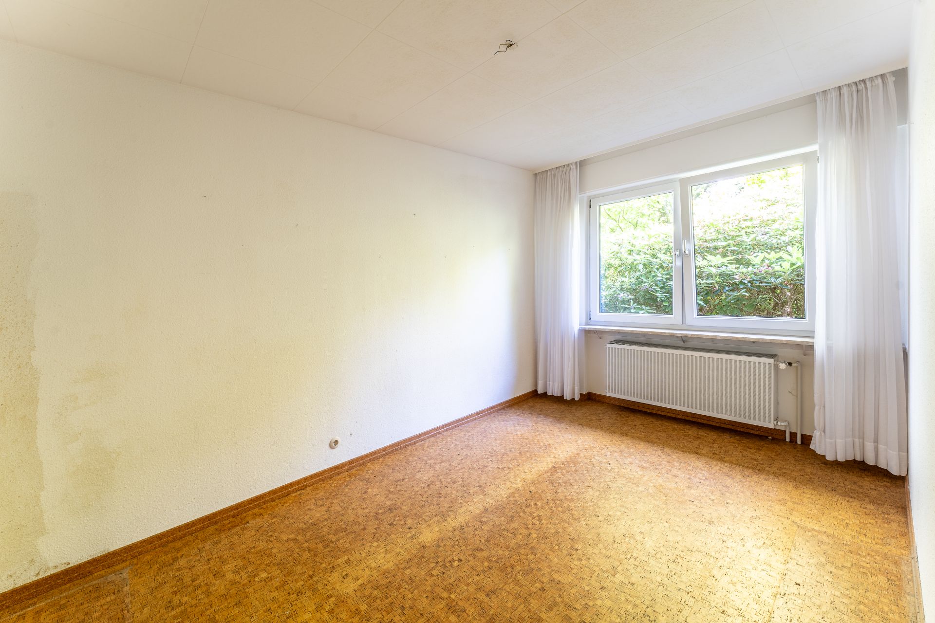 Leeres Zimmer mit großem Fenster, weißen Vorhängen und Heizkörper unter dem Fenster.