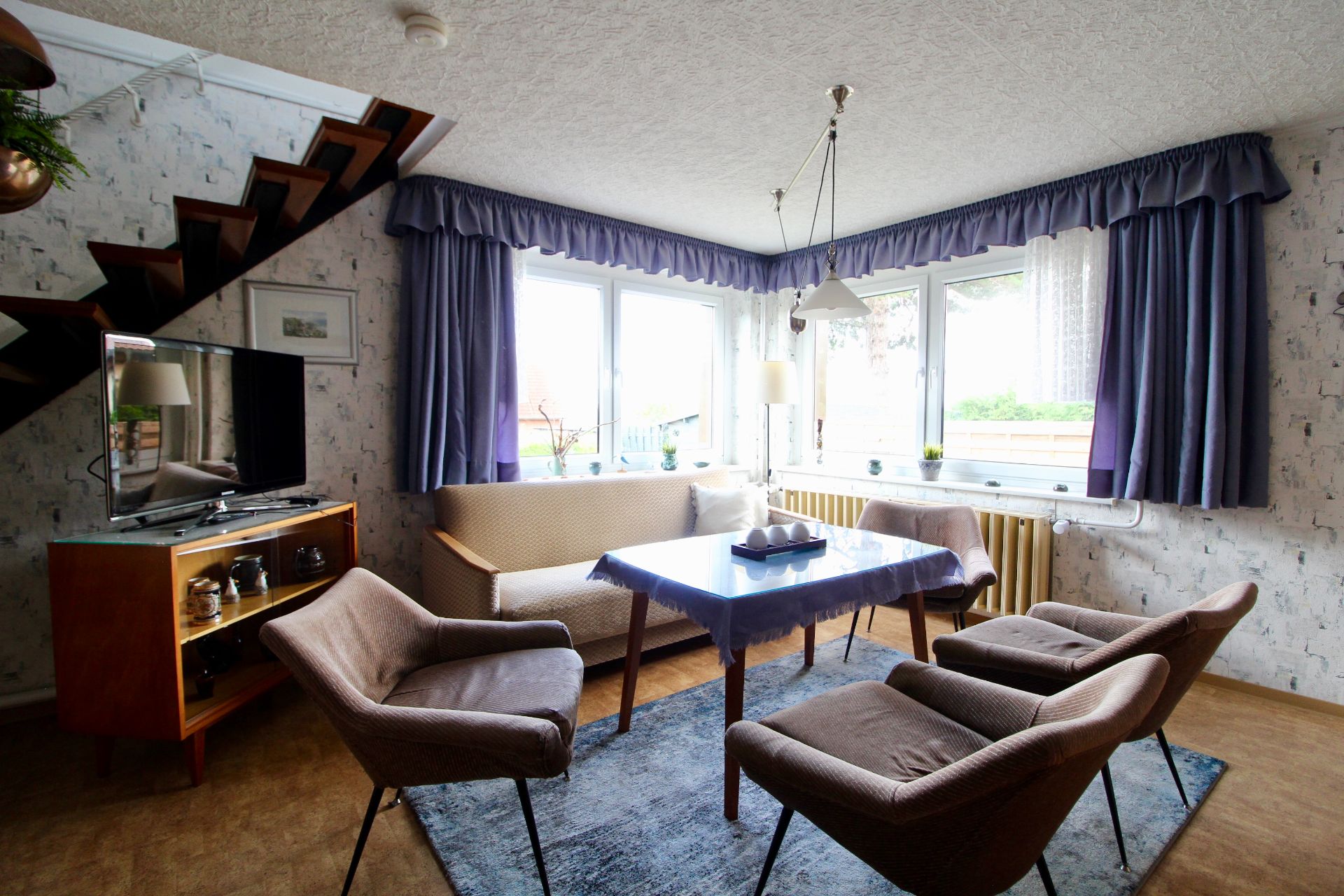 Wohnzimmer mit Sofa, vier Sesseln, Tisch, Fernseher, Treppe und großen Fenstern mit lila Vorhängen.