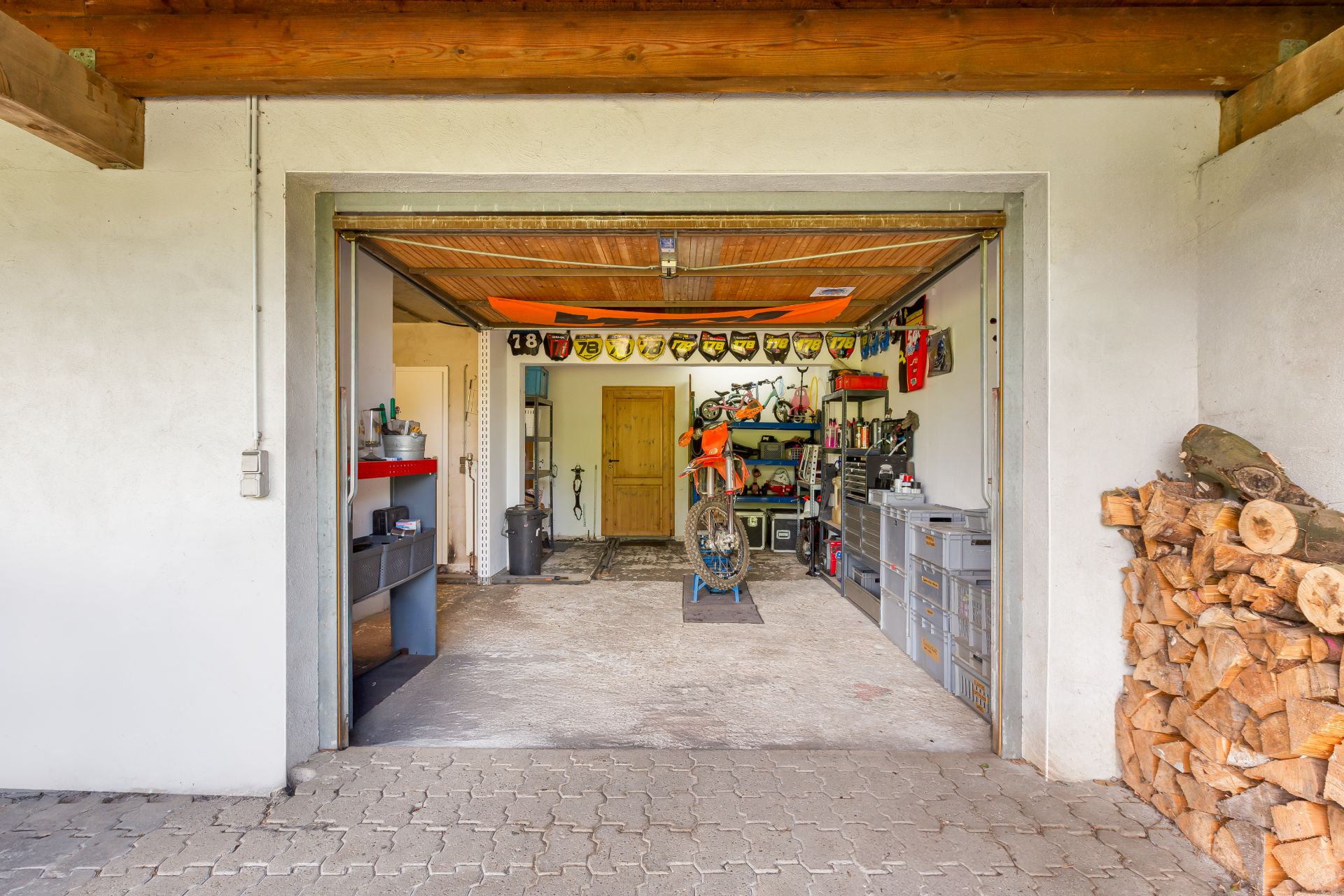 Geöffnete Garage mit Werkbank, Motorrad, Werkzeugen und gestapeltem Brennholz neben dem Eingang.