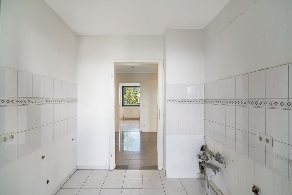 Leerer, gefliester Raum mit Anschlüssen, offene Tür führt in ein weiteres Zimmer mit Fenster.