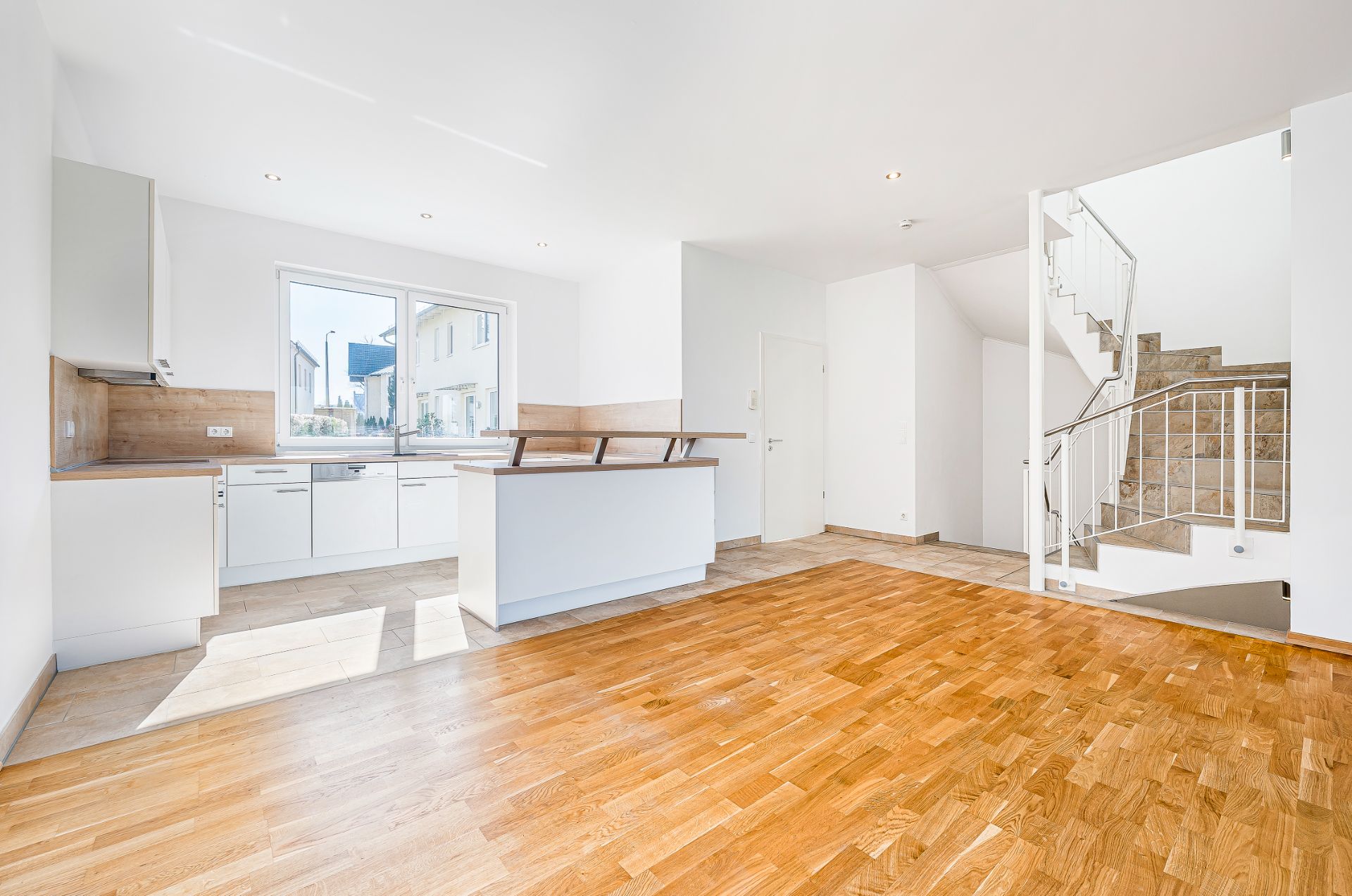 Heller Wohnbereich mit offener Küche, Parkettboden und moderner Treppe im Hintergrund.