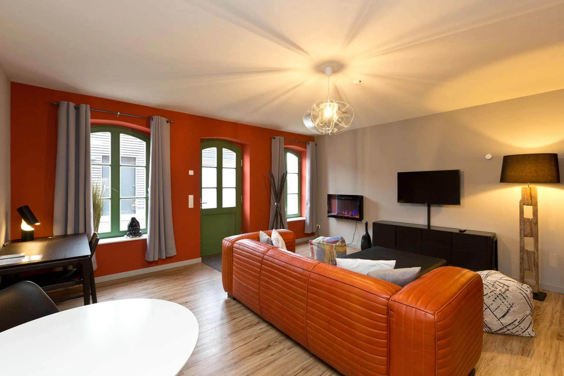 Modernes Wohnzimmer mit orangefarbener Couch, Schreibtisch, TV, Stehlampe und großen Fenstern.