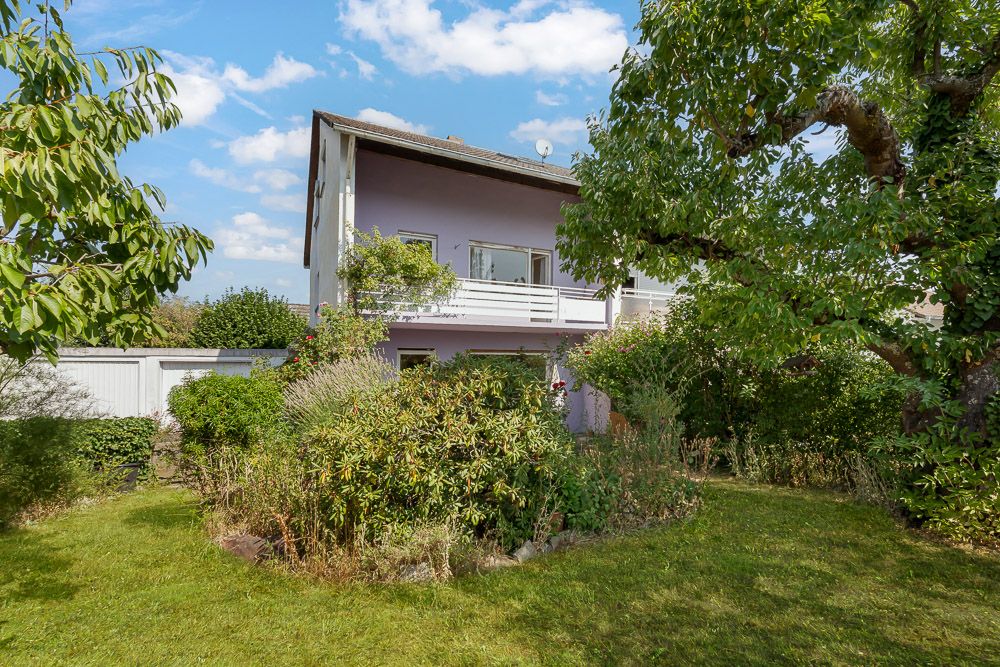 Zweistöckiges Haus mit Balkon und großem Garten, umgeben von Bäumen und Sträuchern.