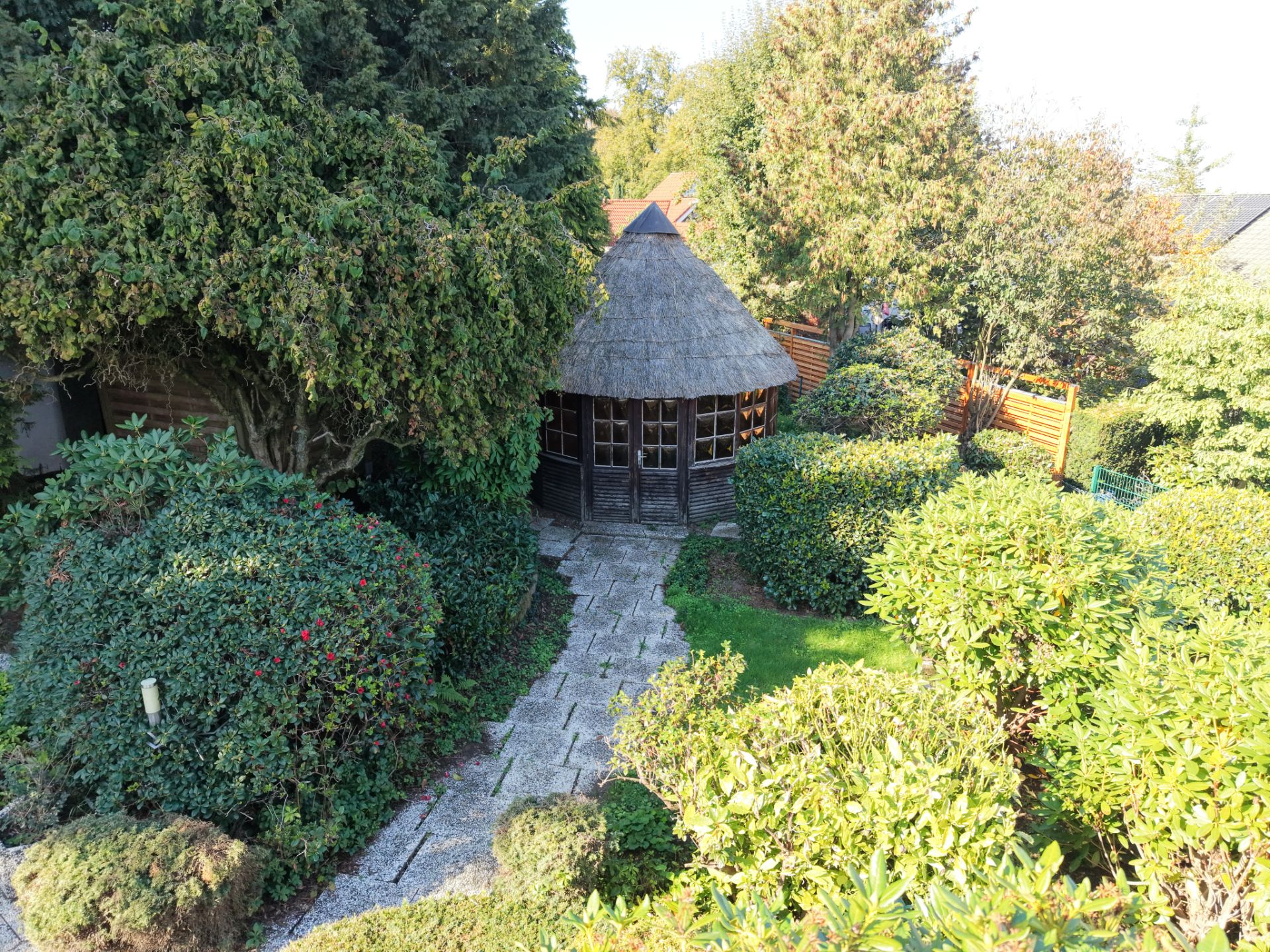 Garten mit Gehweg und rundem Pavillon aus Holz, umgeben von Sträuchern und Bäumen.
