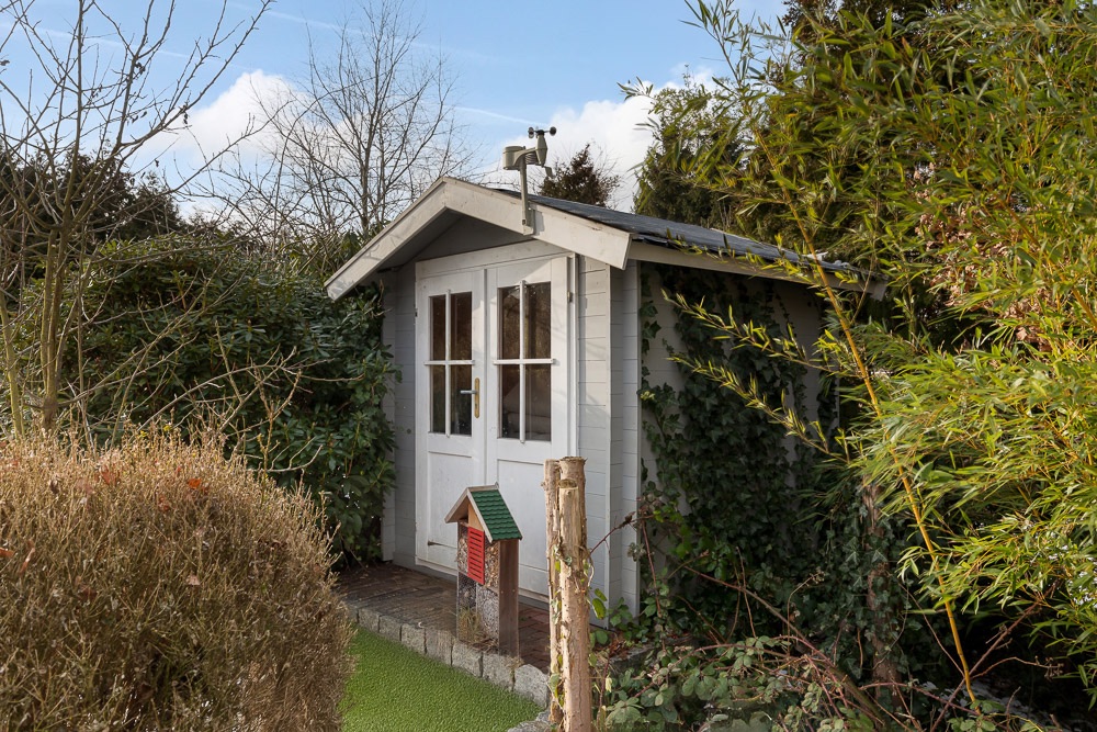 Kleines Gartenhaus mit weißen Türen, umgeben von Büschen und Bäumen im Garten.