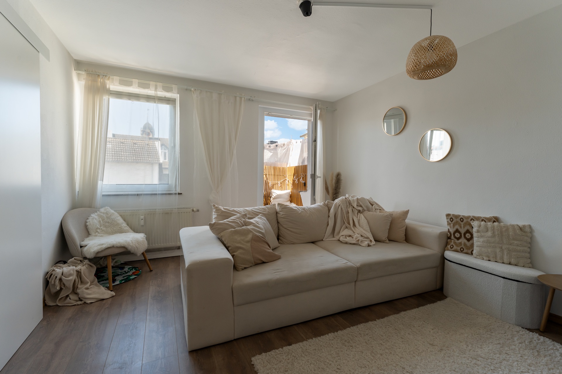 Wohnzimmer mit heller Couch, Kissen, zwei Fenstern, Holzfußboden und drei runden Spiegeln an der Wand.