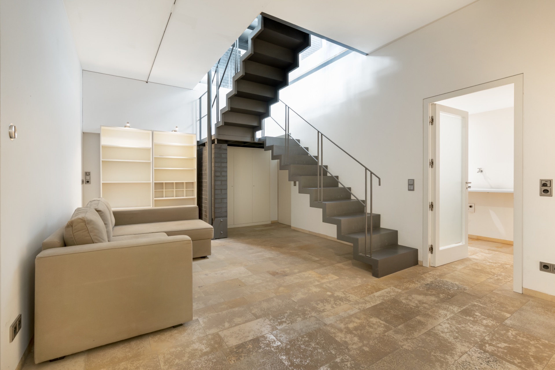 Helles Wohnzimmer mit beige Sofa, Regalwand und moderner Treppe, Tür zum Nebenraum.